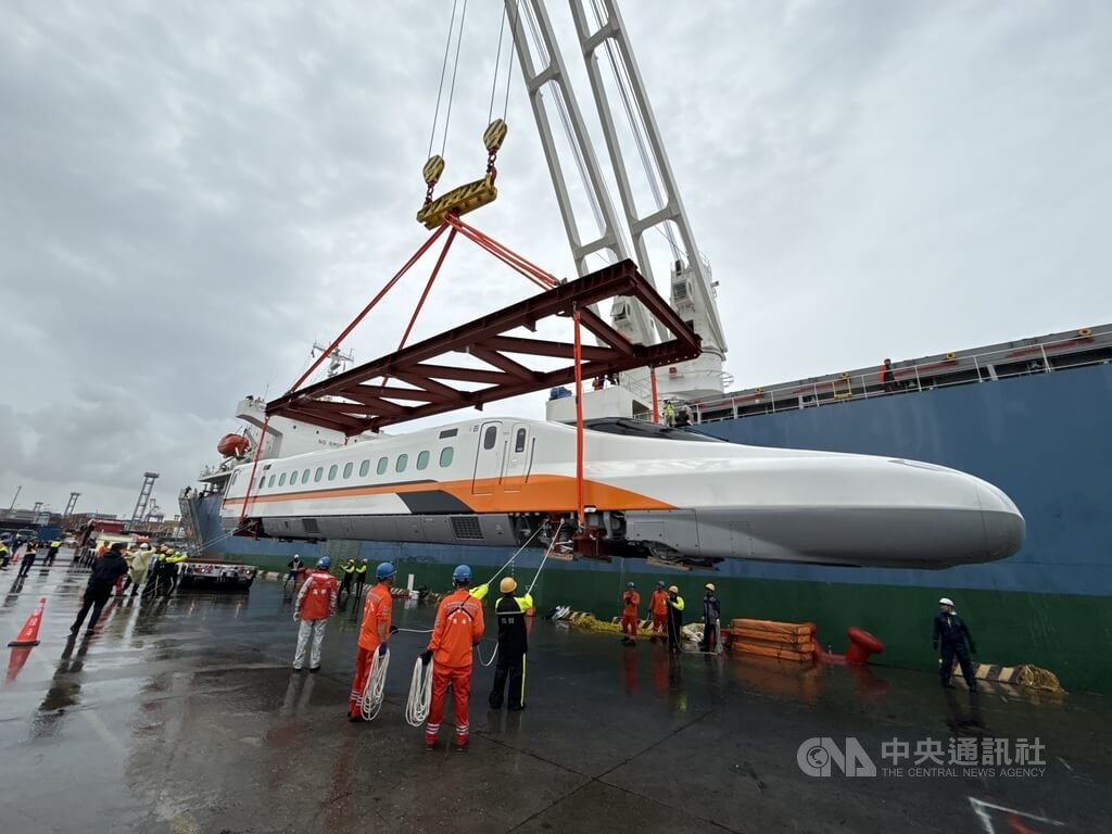 台湾白、橙、黑三色的N700ST高铁列车由吊架悬在货轮旁，工作人员在高雄港被雨水浸湿的码头上引导车辆落位。