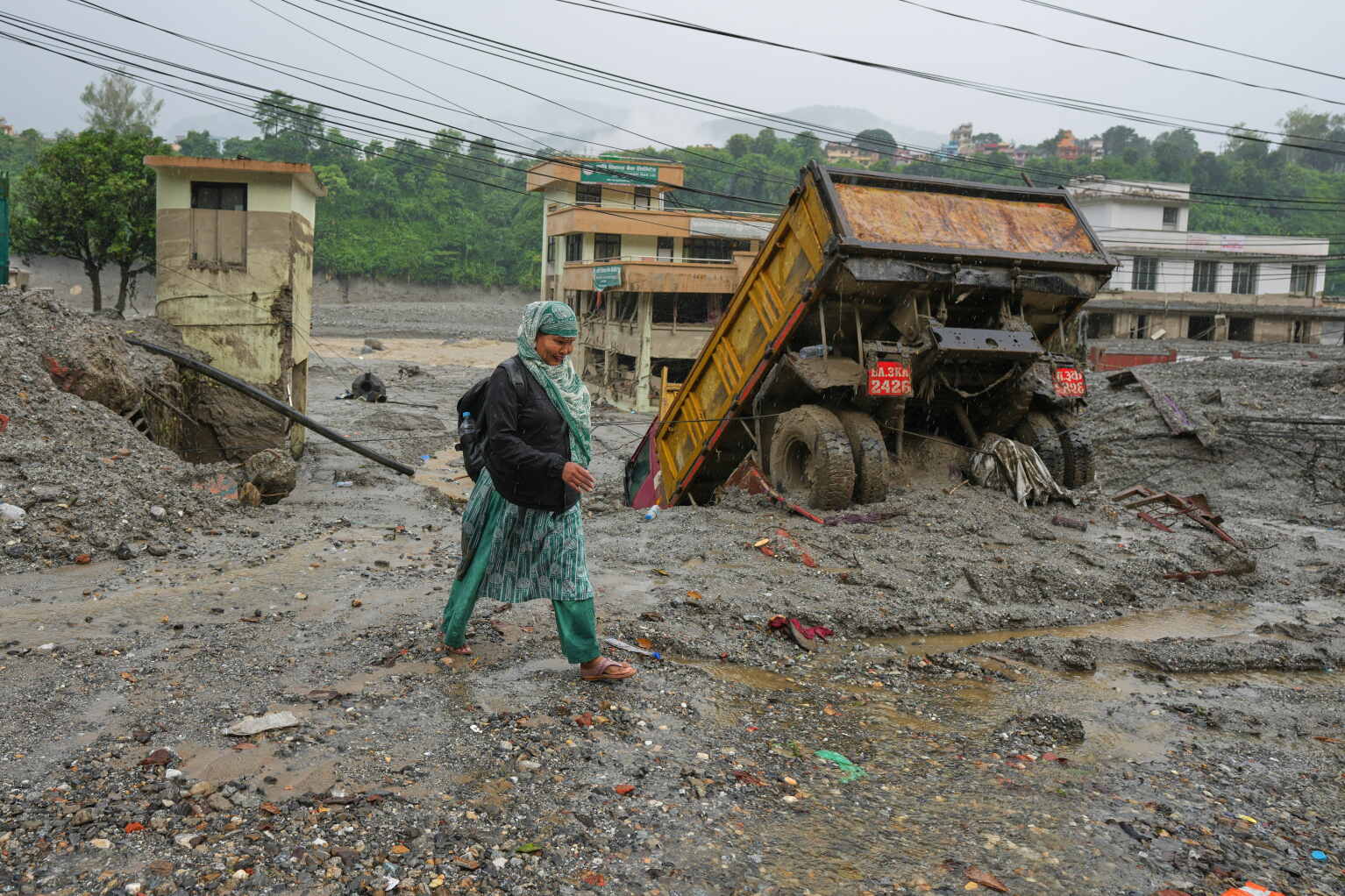 A woman walks across grey flood mud beside a half-buried dump truck and damaged buildings in Nuwakot, Nepal.
