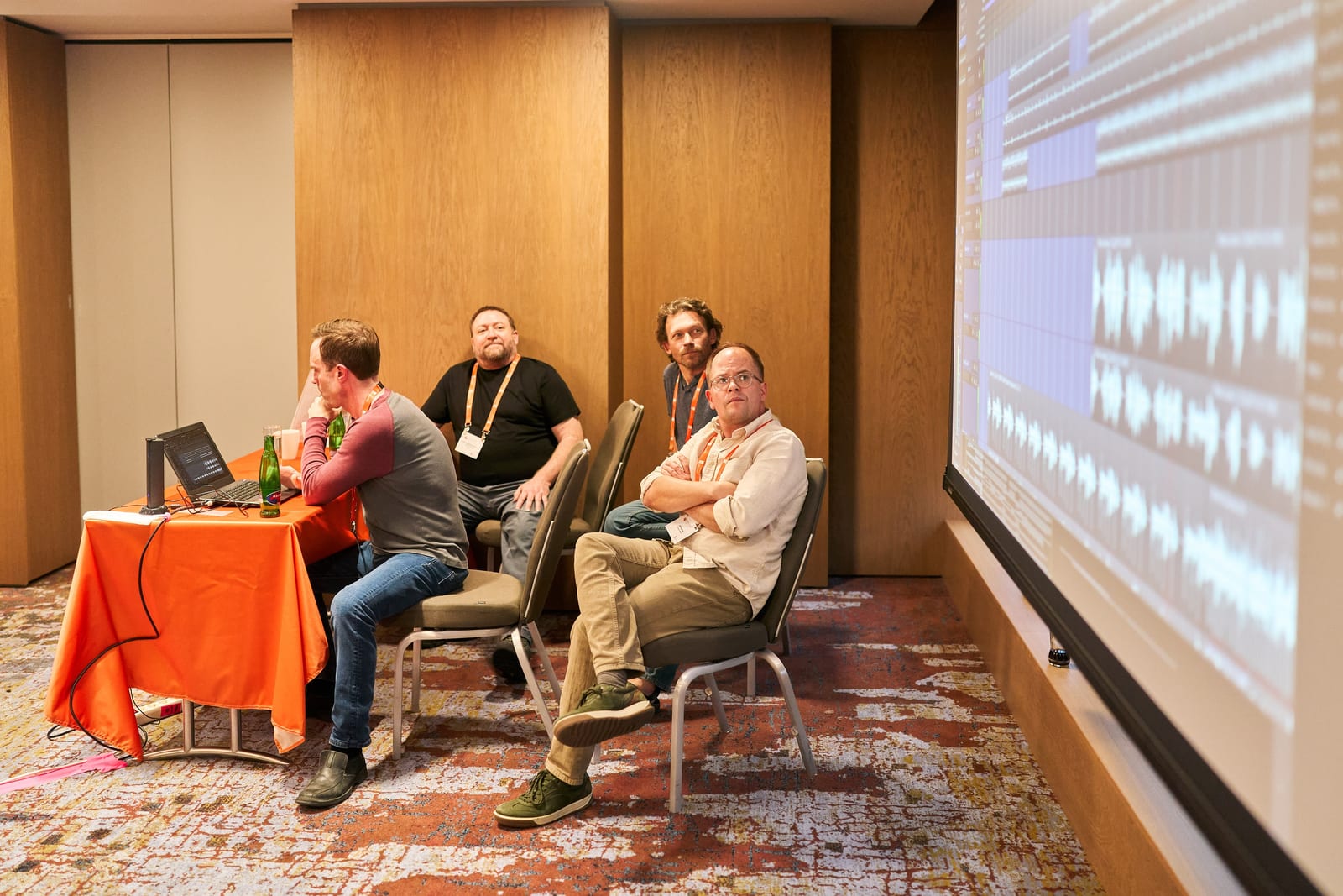 Four musicians sit around a laptop during an Ardour workshop while multitrack audio waveforms fill a projection screen beside them.