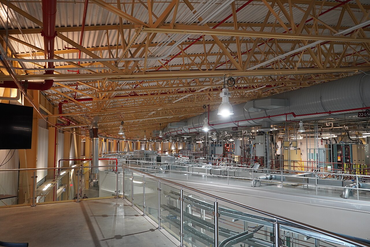 A view from an upper walkway across the circular NSLS-II experimental floor, showing silver beamline enclosures, pipes, equipment cabinets, and overhead trusses.