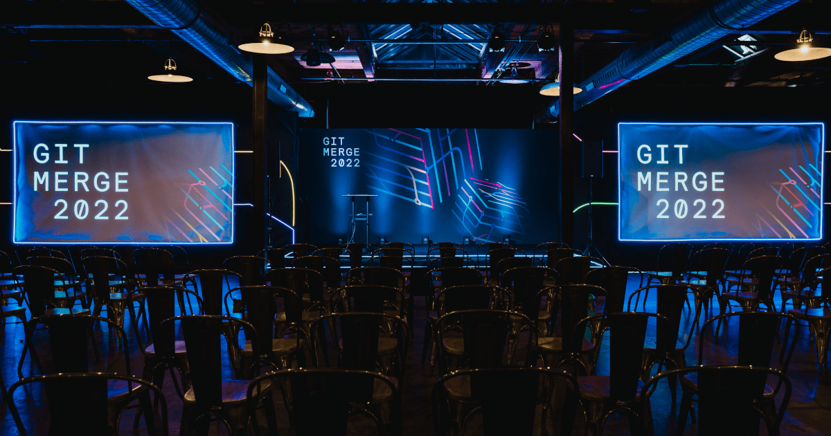 Empty rows of chairs face three blue-lit screens reading Git Merge 2022 at the conference venue in Chicago.