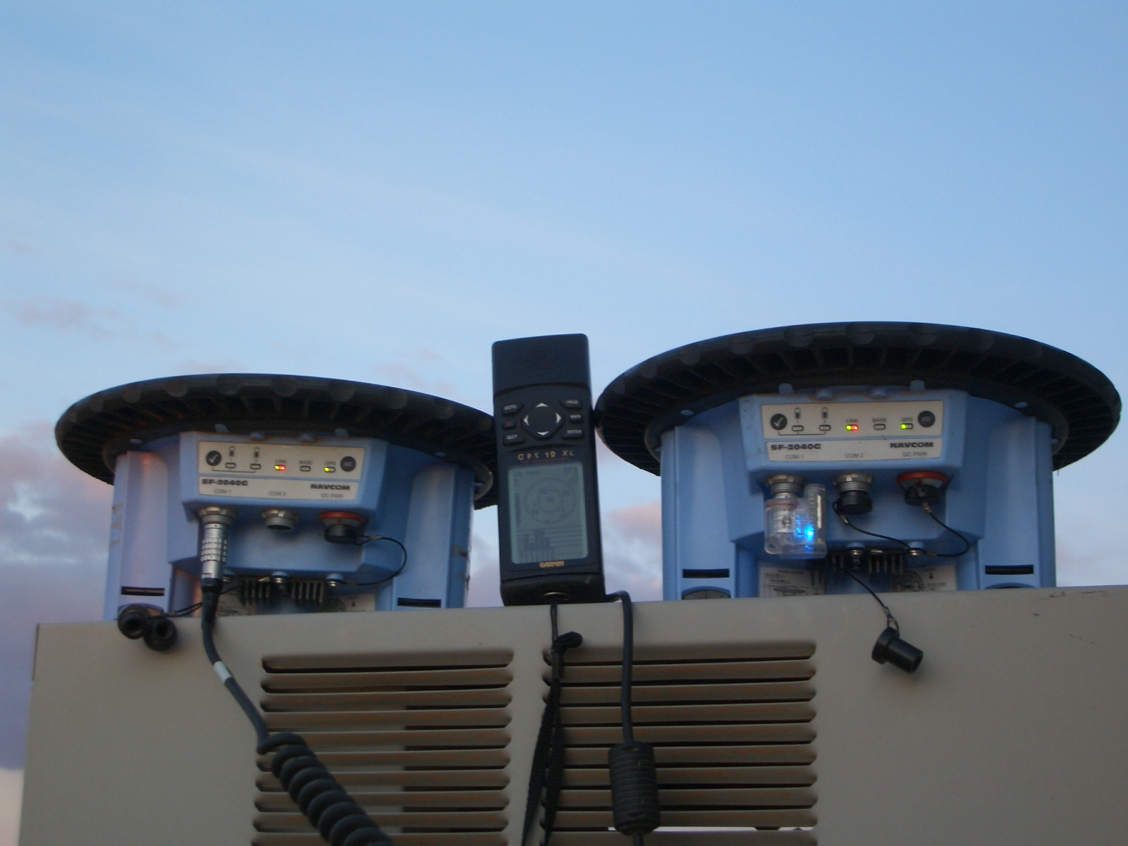 Two Navcom GNSS receivers flank a Garmin handheld, with cables and a glowing Bluetooth serial adapter visible against an evening sky.