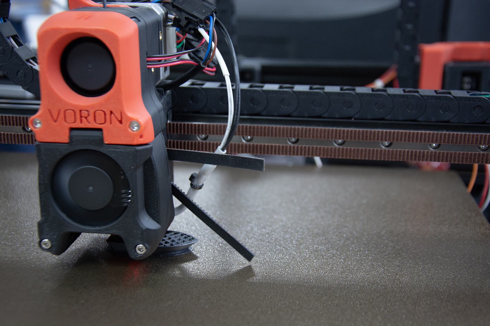 一台 Voron 3D 打印机正在热床上铺设耗材，近处可见橙色工具头、皮带、拖链和机架。