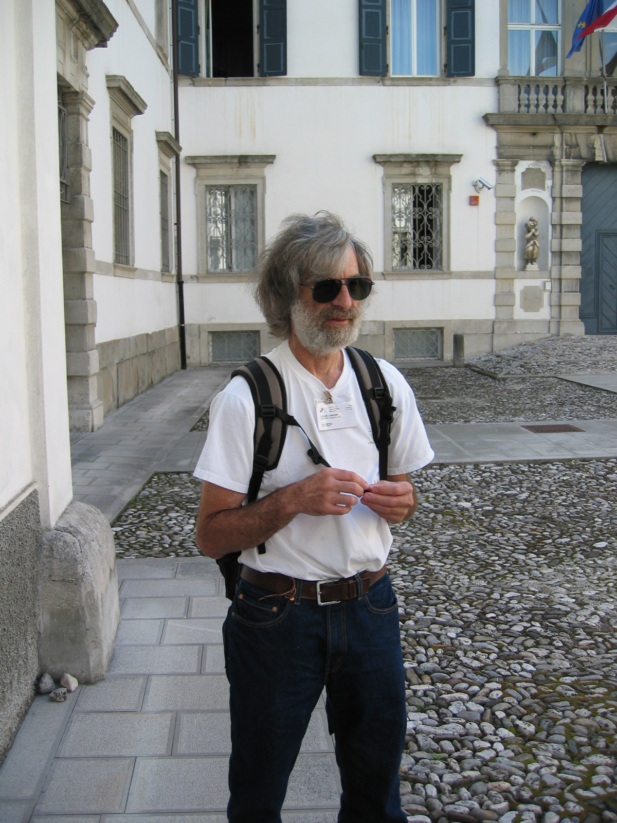 Leslie Lamport 戴着墨镜和会议证件，站在意大利乌迪内 IFIP 会场的一处石砌庭院里。