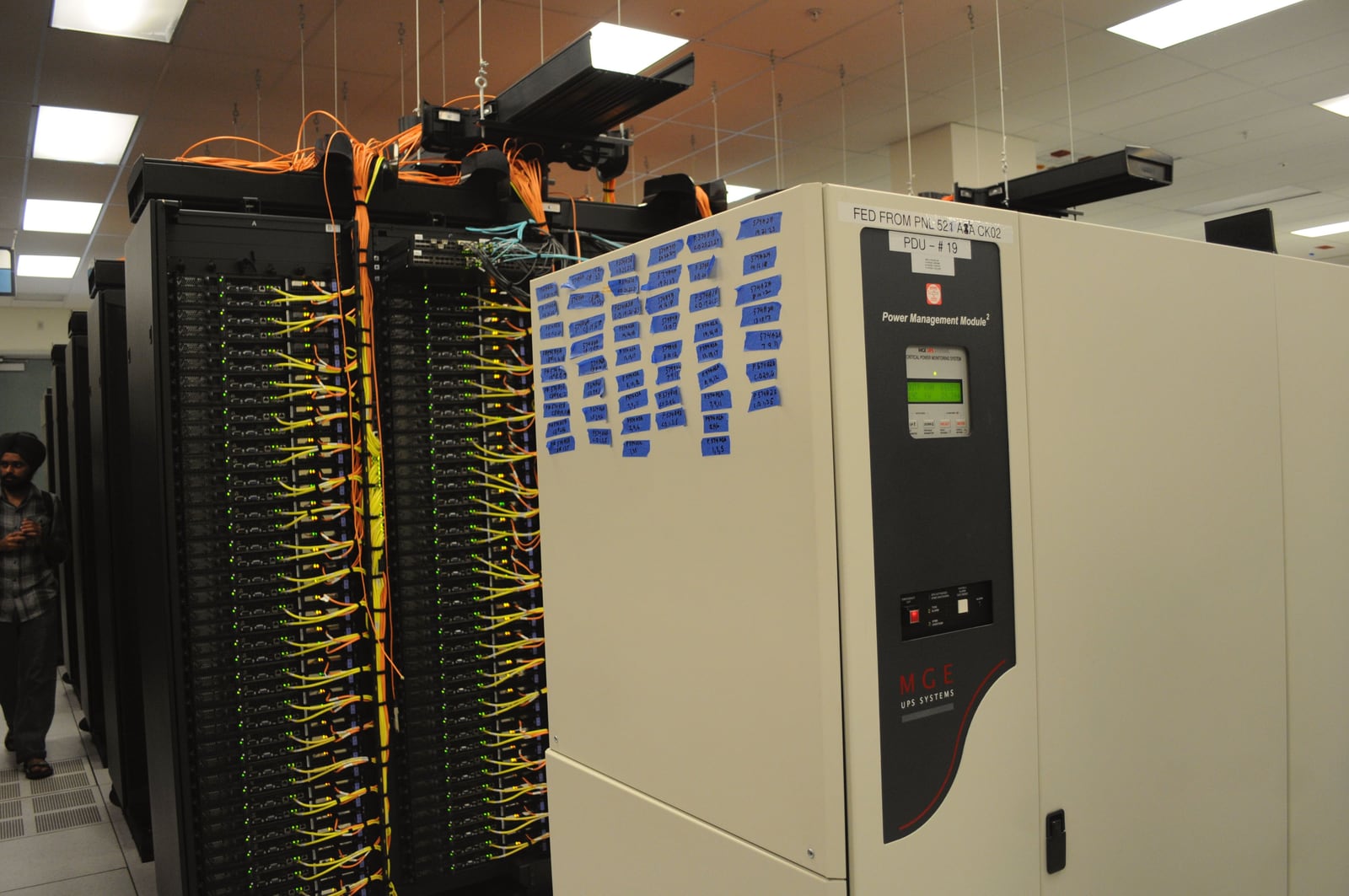 A UPS power-management cabinet stands beside rows of network racks and blue cabling in the NERSC data center.