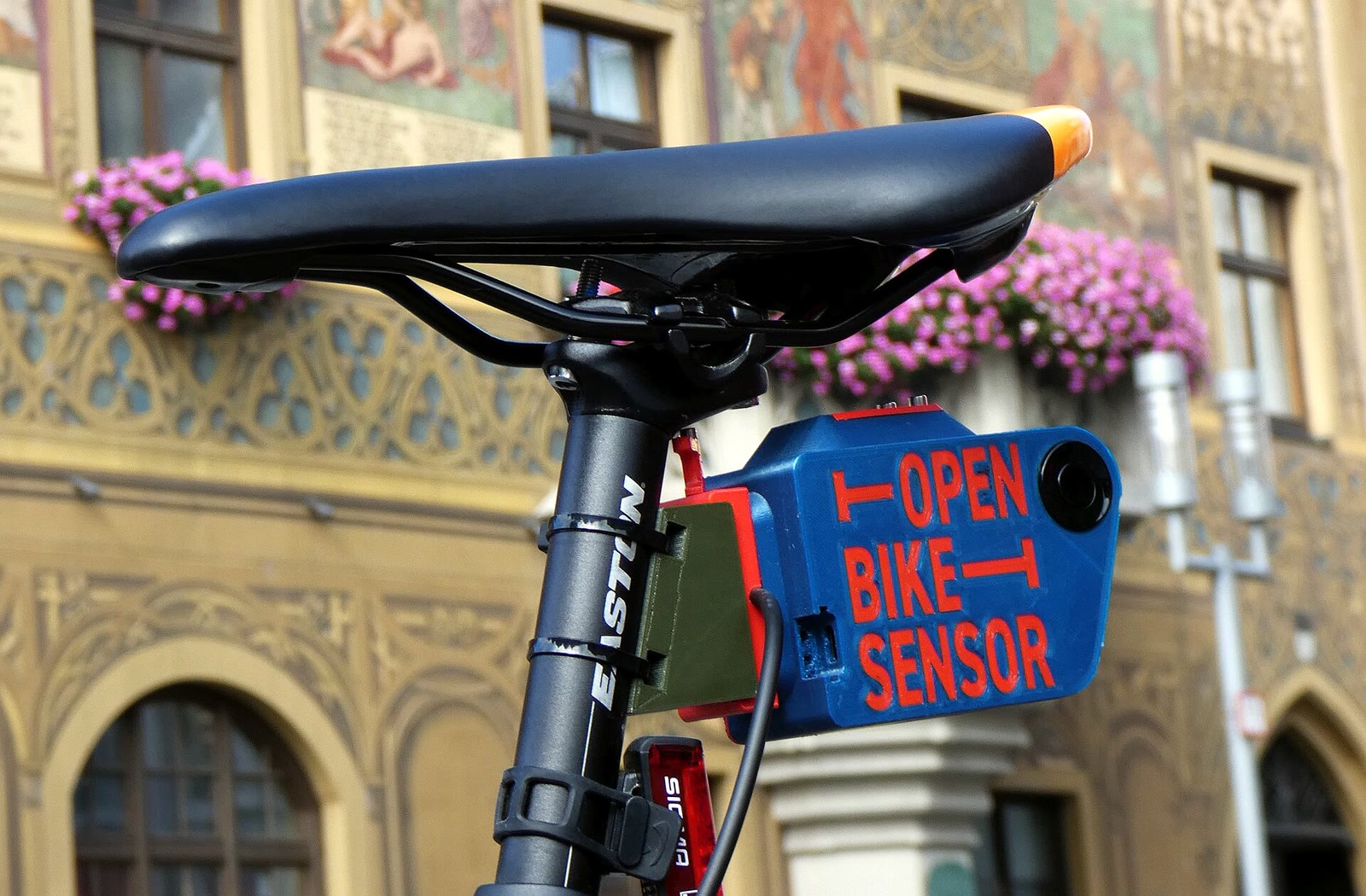 A blue and red OpenBikeSensor with its round ultrasonic transducer mounted beneath a bicycle saddle in front of Ulm City Hall.