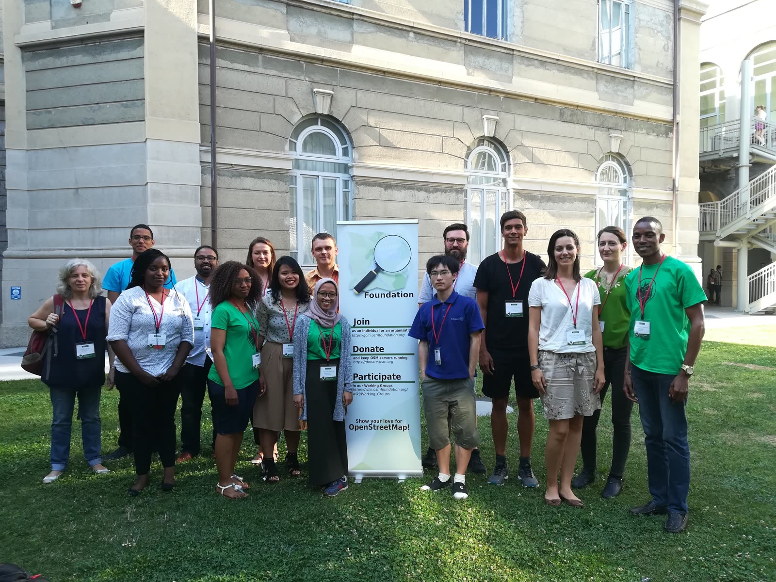 State of the Map 2018 scholarship recipients stand beside an OpenStreetMap Foundation banner in a Milan courtyard.