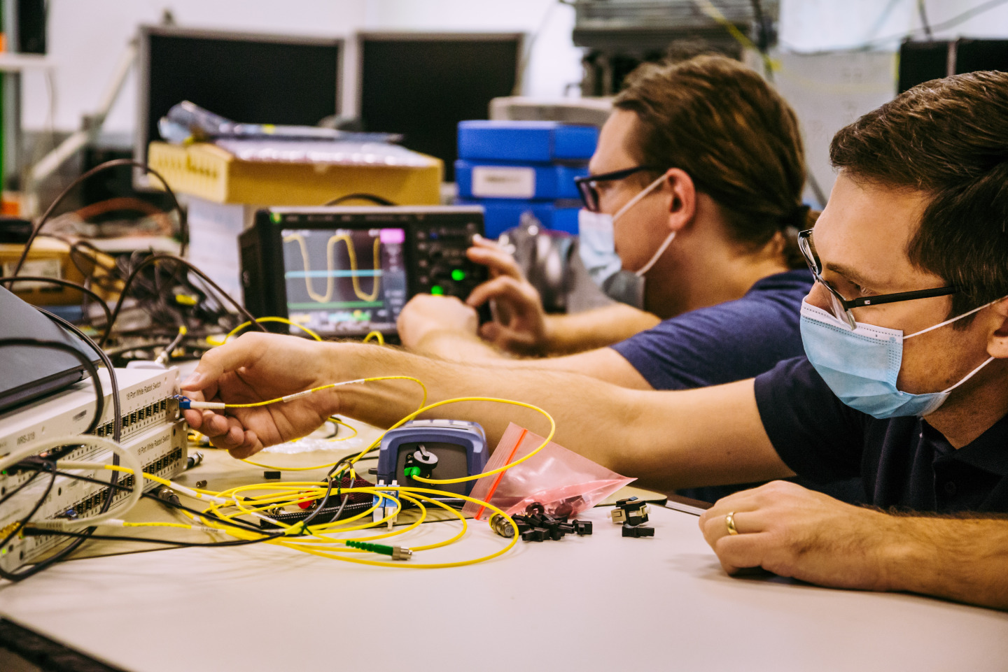 Two engineers testing White Rabbit network switches and yellow optical-fiber links on a CERN laboratory bench.