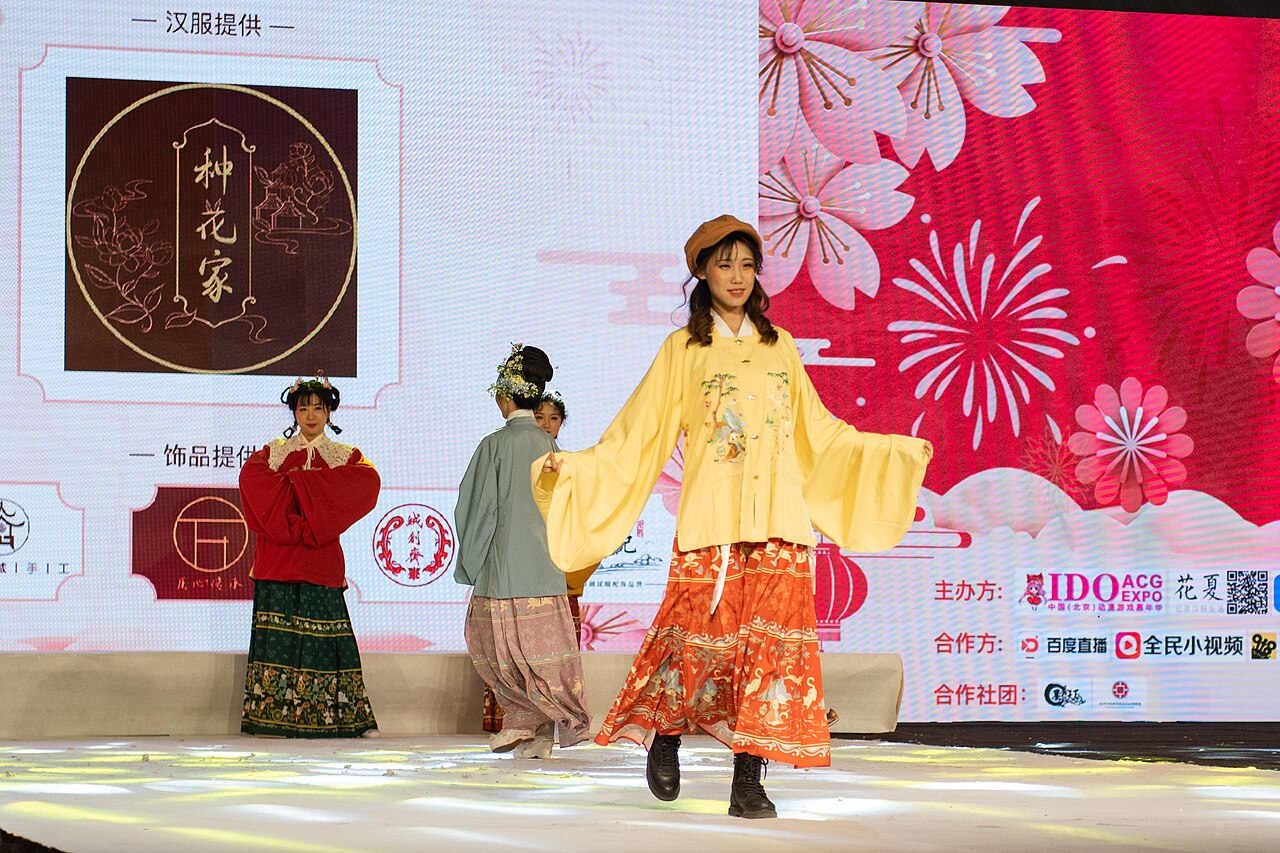 Models in colorful Hanfu-inspired outfits walk an exhibition stage, pairing long skirts and wide sleeves with modern shoes and a beret.