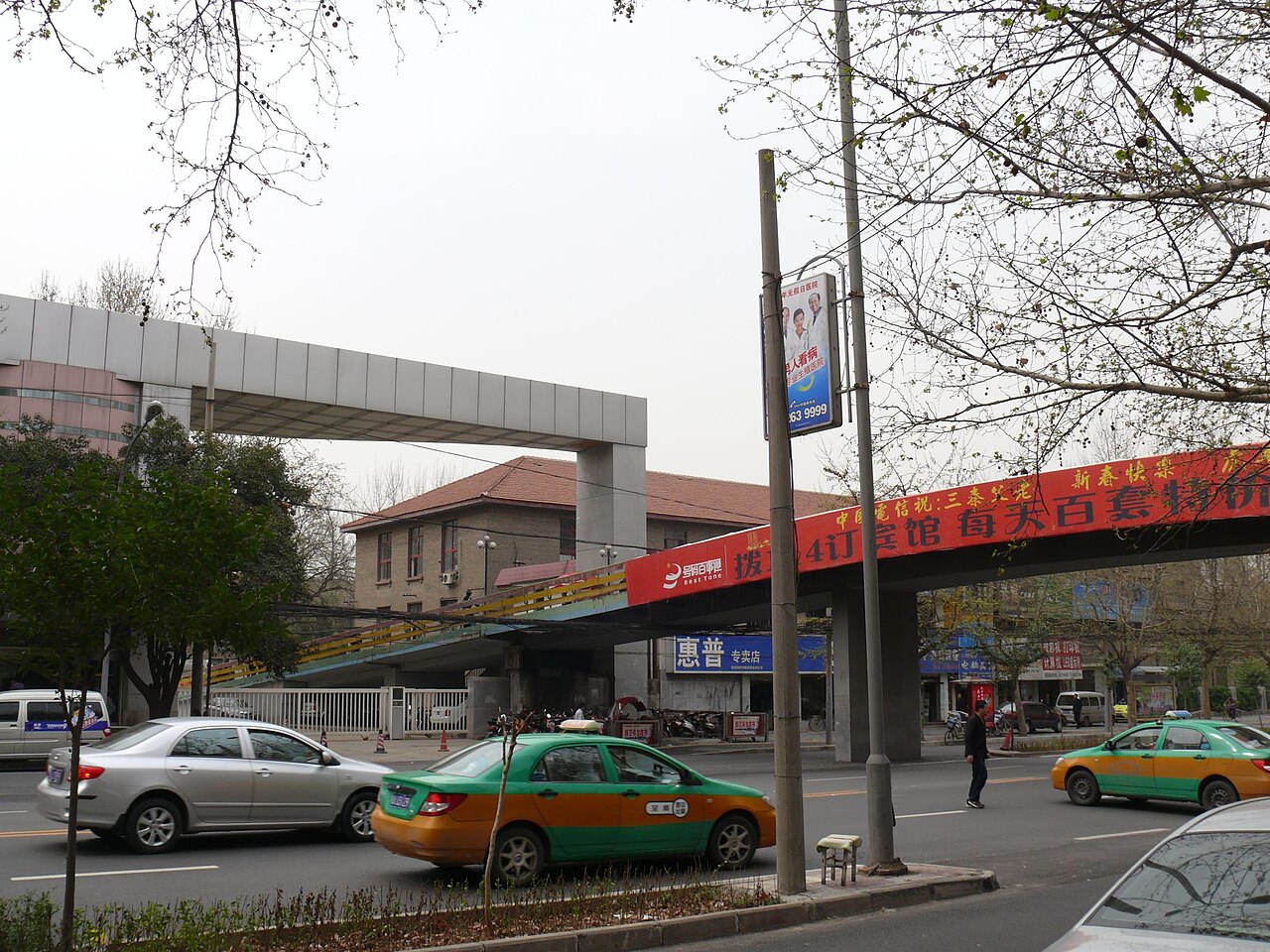 Green and yellow taxis pass an entrance to Xi’an Jiaotong University beneath a pedestrian bridge and roadside trees.