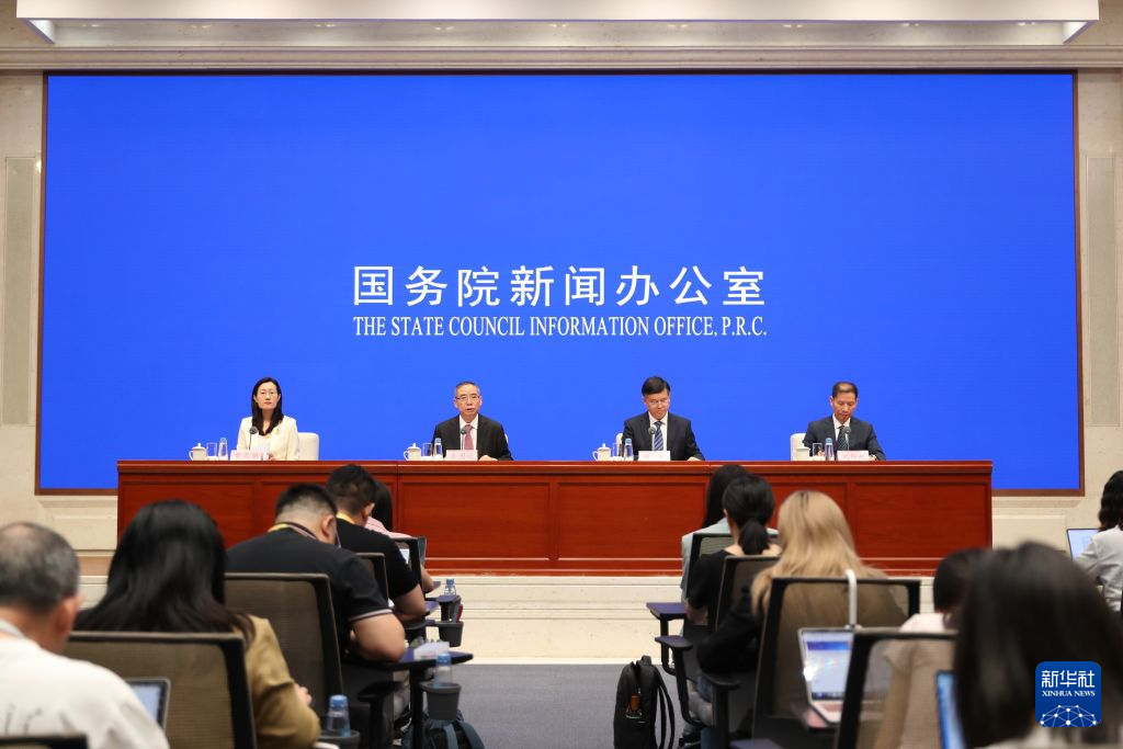 在北京国务院新闻办公室的一场新闻发布会上，四名官员坐在麦克风后方，画面前方的记者正在工作。