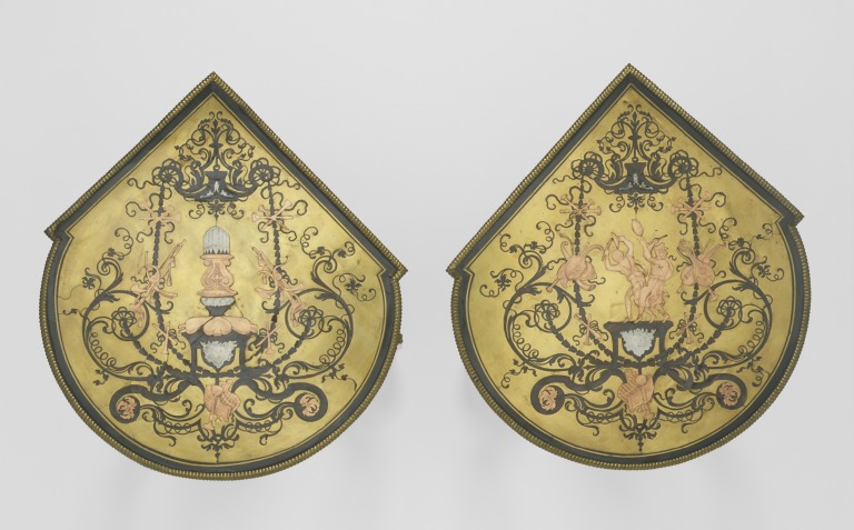 Two shaped table tops photographed from above, with dark scrolling ornament and pale metal details set into golden brass grounds.