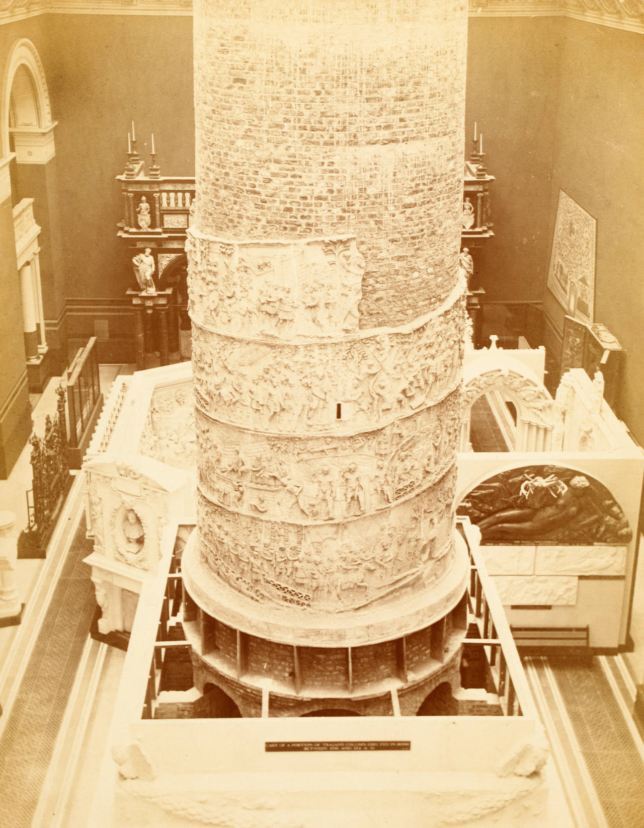 Sepia photograph of Trajan's Column under construction in the V&A, with an exposed brick core above bands of plaster relief.