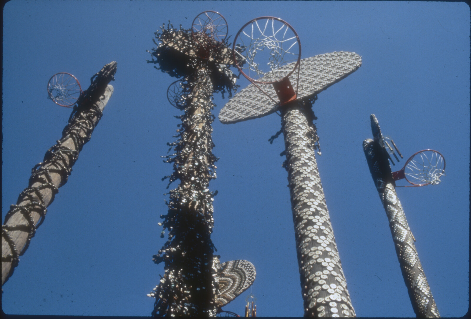 An archival photograph looking up at David Hammons's bottle-cap-covered poles and basketball hoops against a blue sky in Cadman Plaza Park.