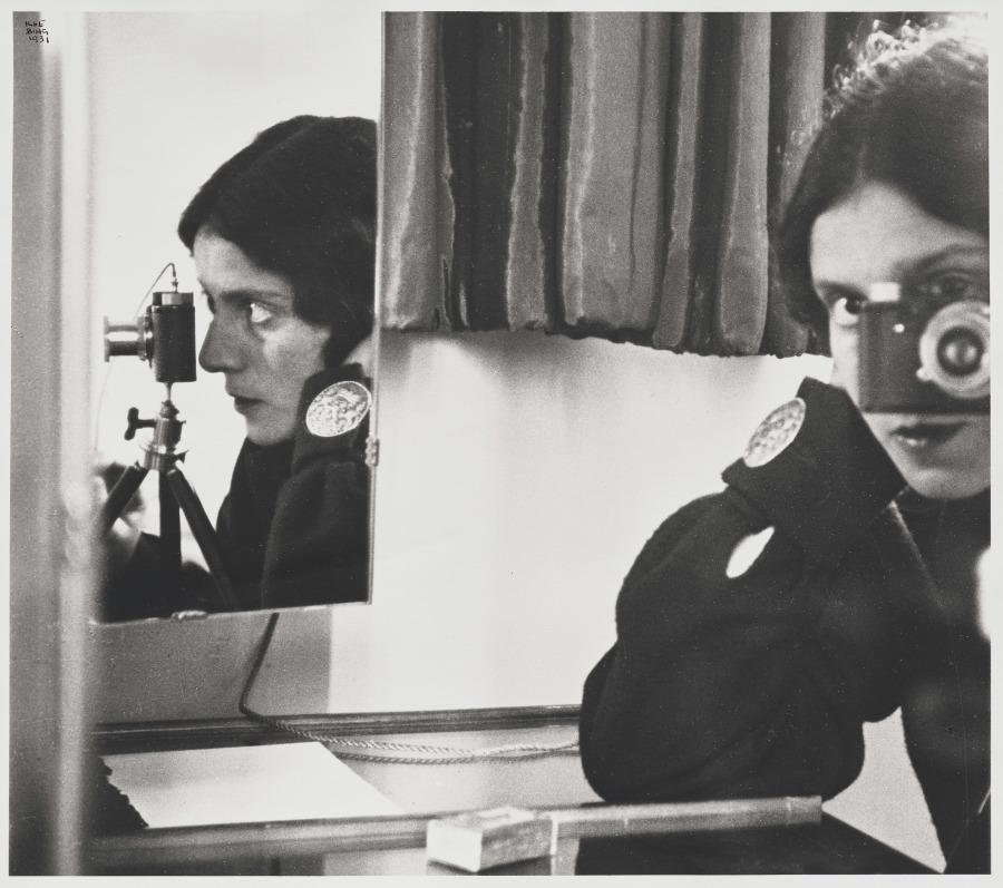 Black-and-white photograph showing Ilse Bing behind a Leica camera, with a second mirror at the left reflecting her face and camera in profile.