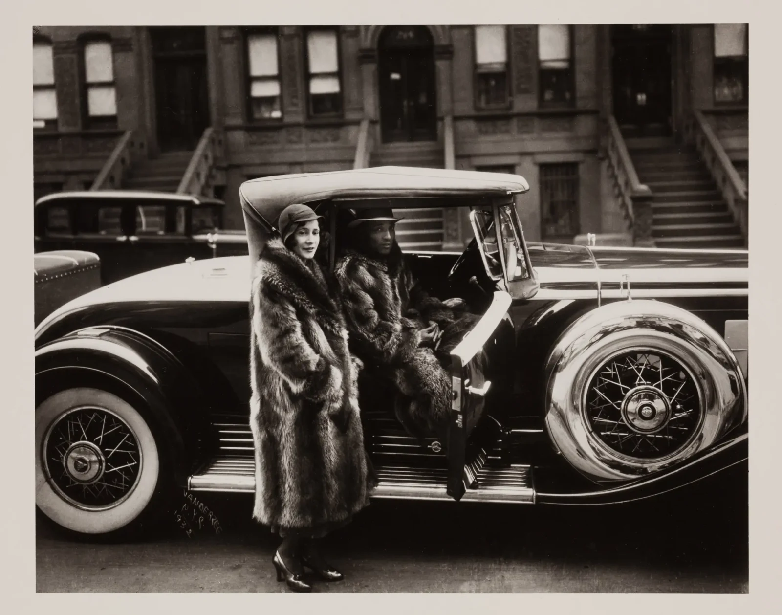 James Van Der Zee's black-and-white photograph of a man seated in a convertible and a woman standing beside it, both wearing long fur coats, with Harlem brownstones behind them.