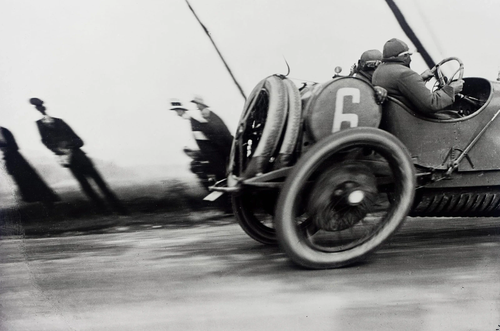 Black-and-white photograph of a racing car numbered 6, cut off at the right edge, with an oval wheel and blurred spectators leaning in the opposite direction.