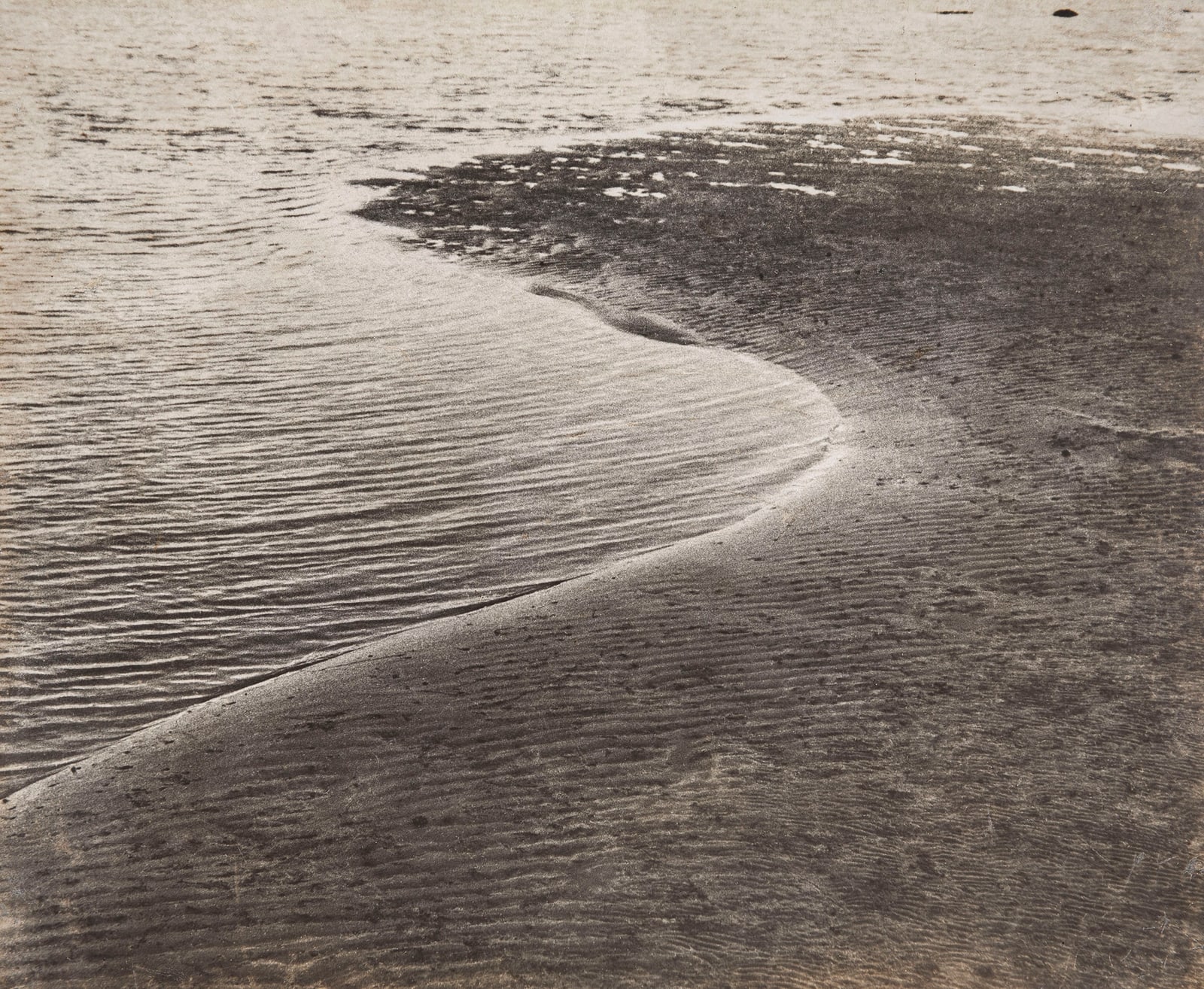 Black-and-white close-up photograph of rippling water curling around a sandbank, creating dense parallel textures and a pale S-shaped shoreline.