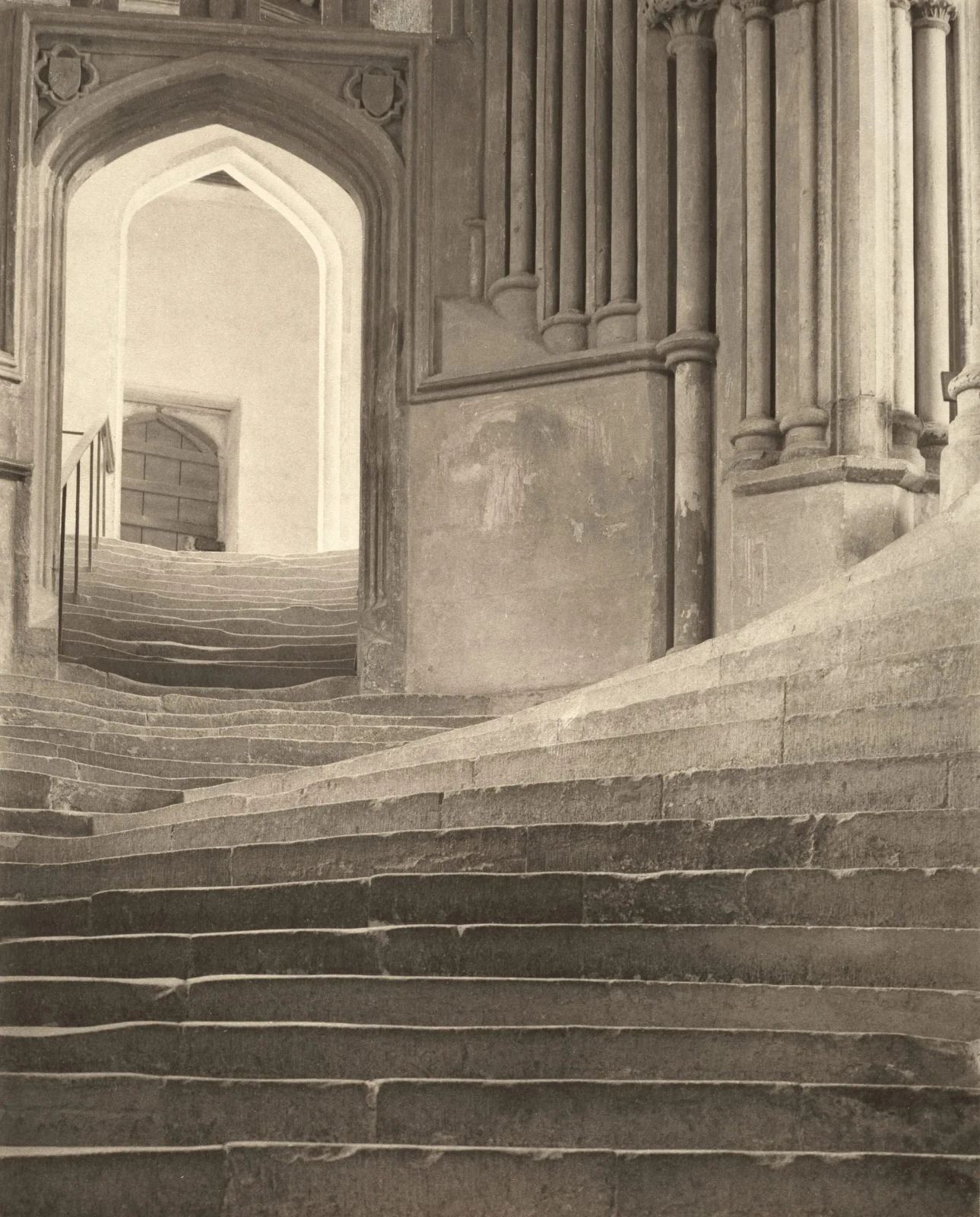 Frederick H. Evans’s warm-toned photograph of worn stone steps rising through pointed arches at Wells Cathedral, with a cluster of columns on the right.
