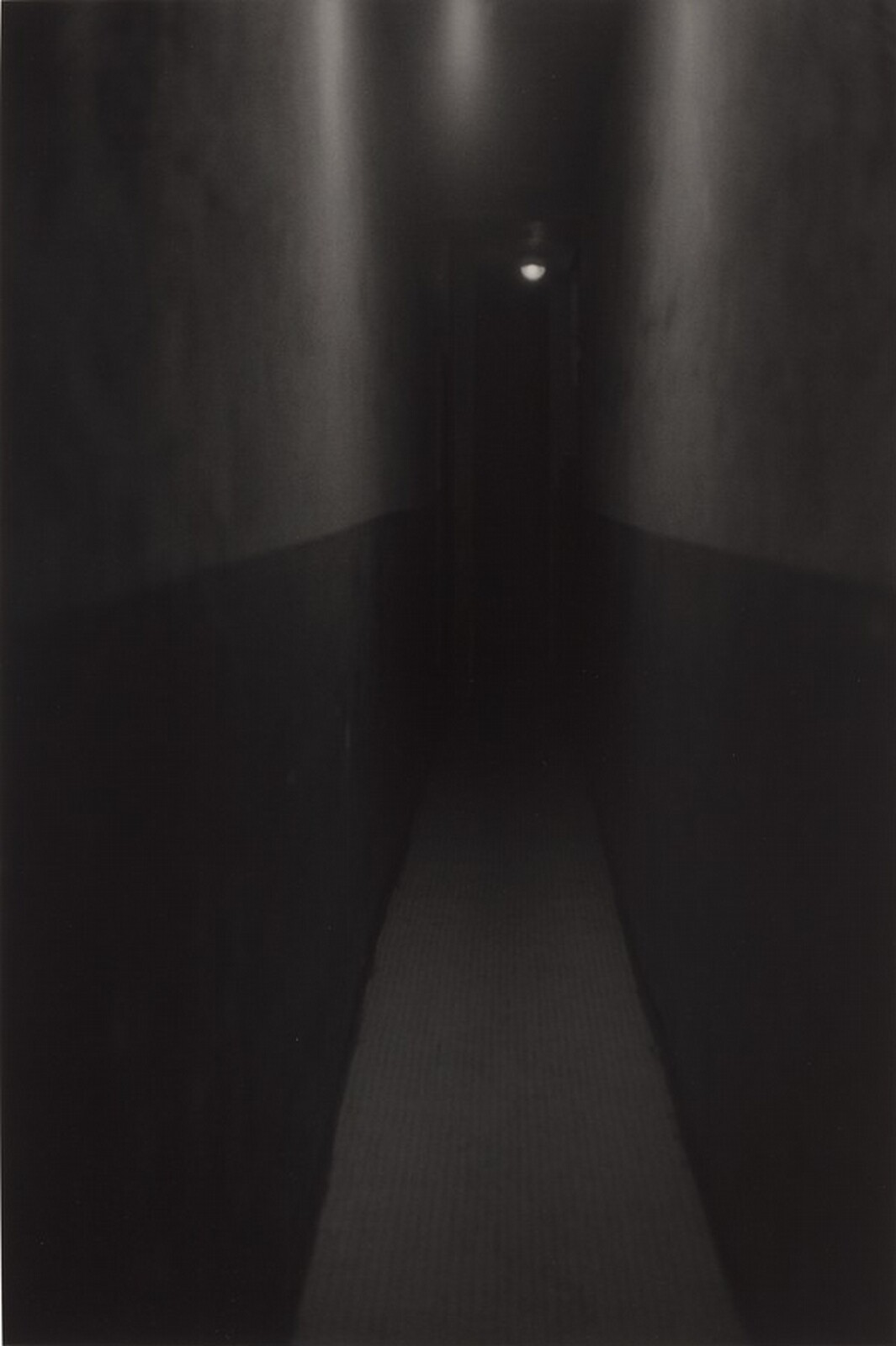 Black-and-white photograph of a narrow, empty hallway, with pale upper walls, dark doorways, and a small ceiling light above a receding floor.