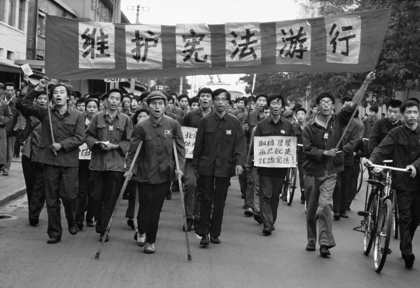 一张黑白档案照片：星星画会的游行者挤满北京街道，头顶的巨幅横幅号召人们参加维护宪法的游行。