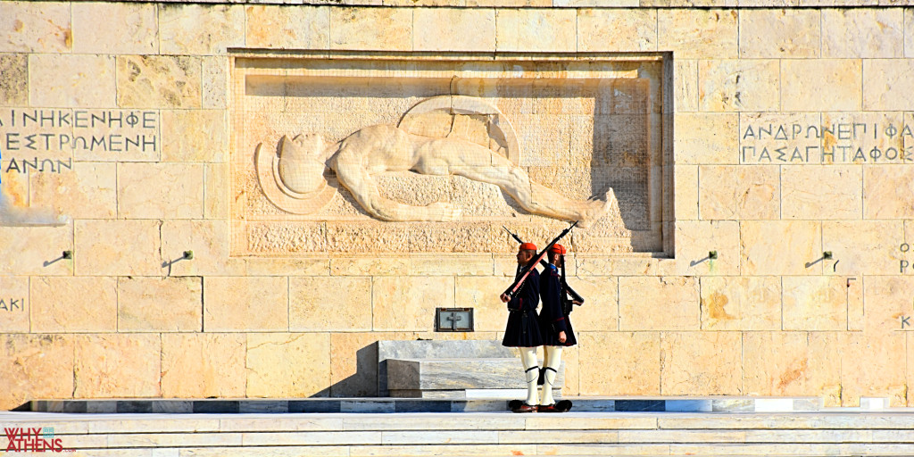 Two Evzones in dark uniforms cross beneath the stone relief of a fallen soldier at Athens’s Tomb of the Unknown Soldier.