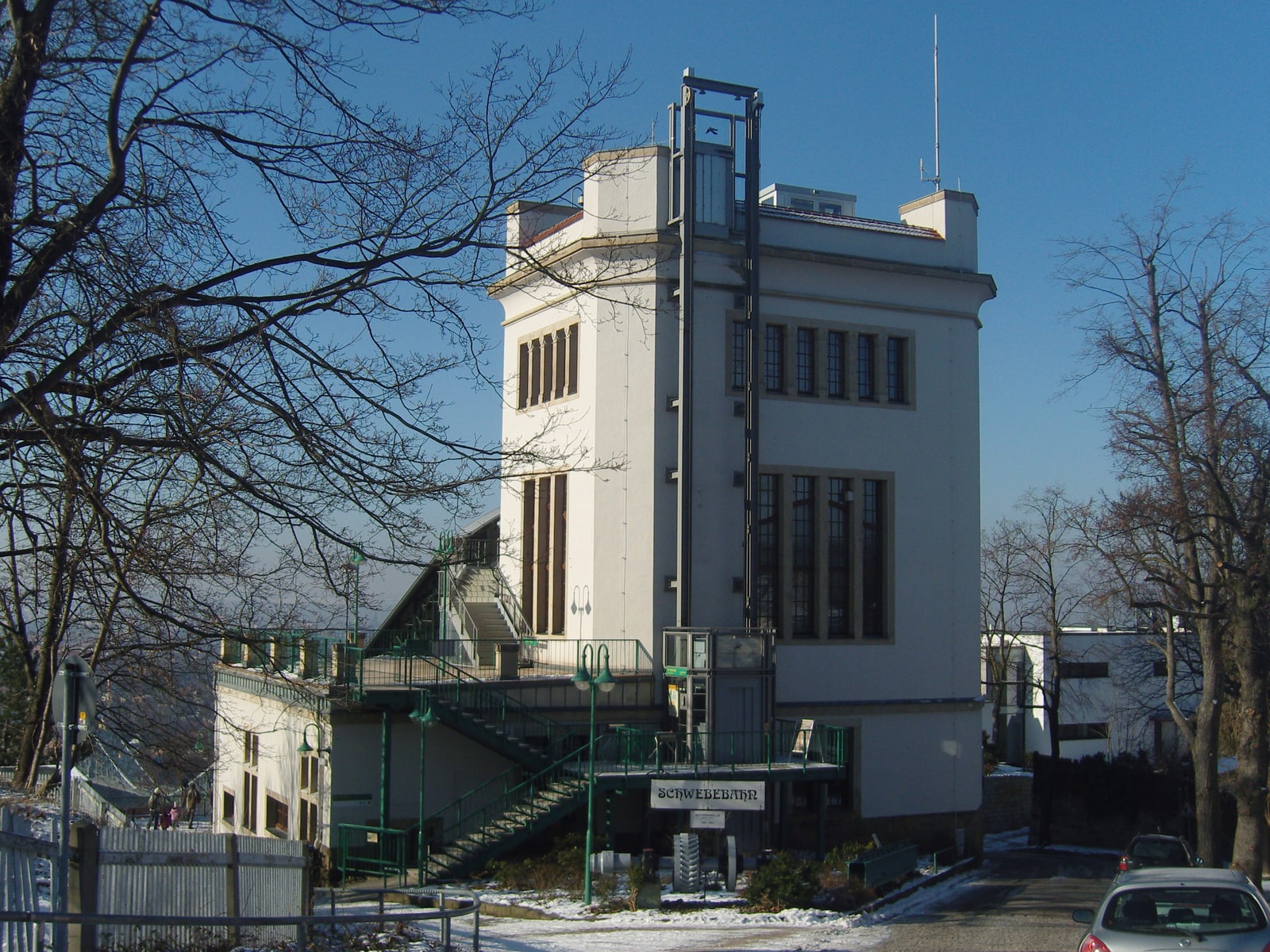 The cream-colored tower of Dresden's Schwebebahn upper station, with an external lift, green railings and a Schwebebahn sign above the entrance.