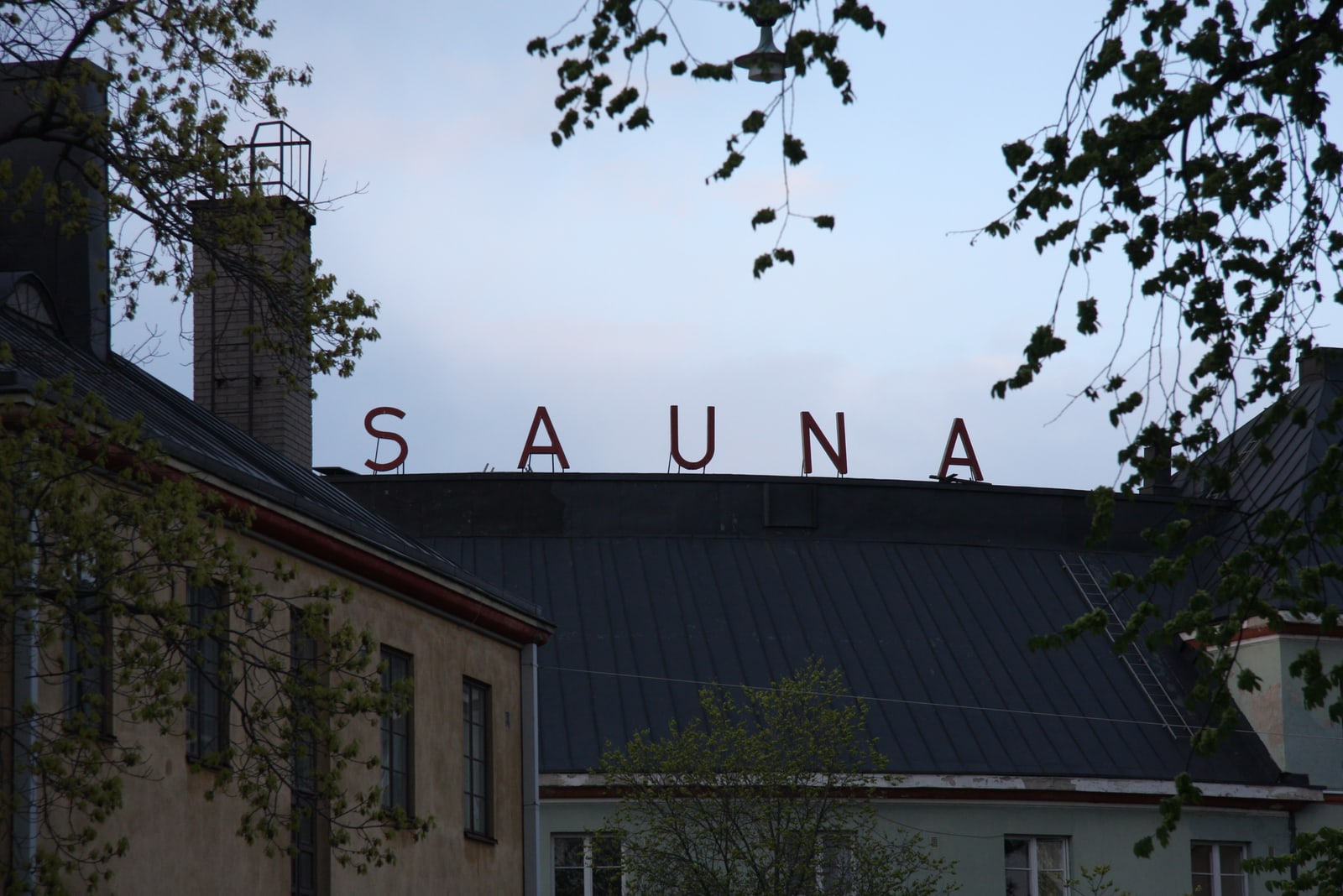 Kotiharjun Sauna's red rooftop SAUNA letters above dark roofs, framed by leafy branches in Helsinki.
