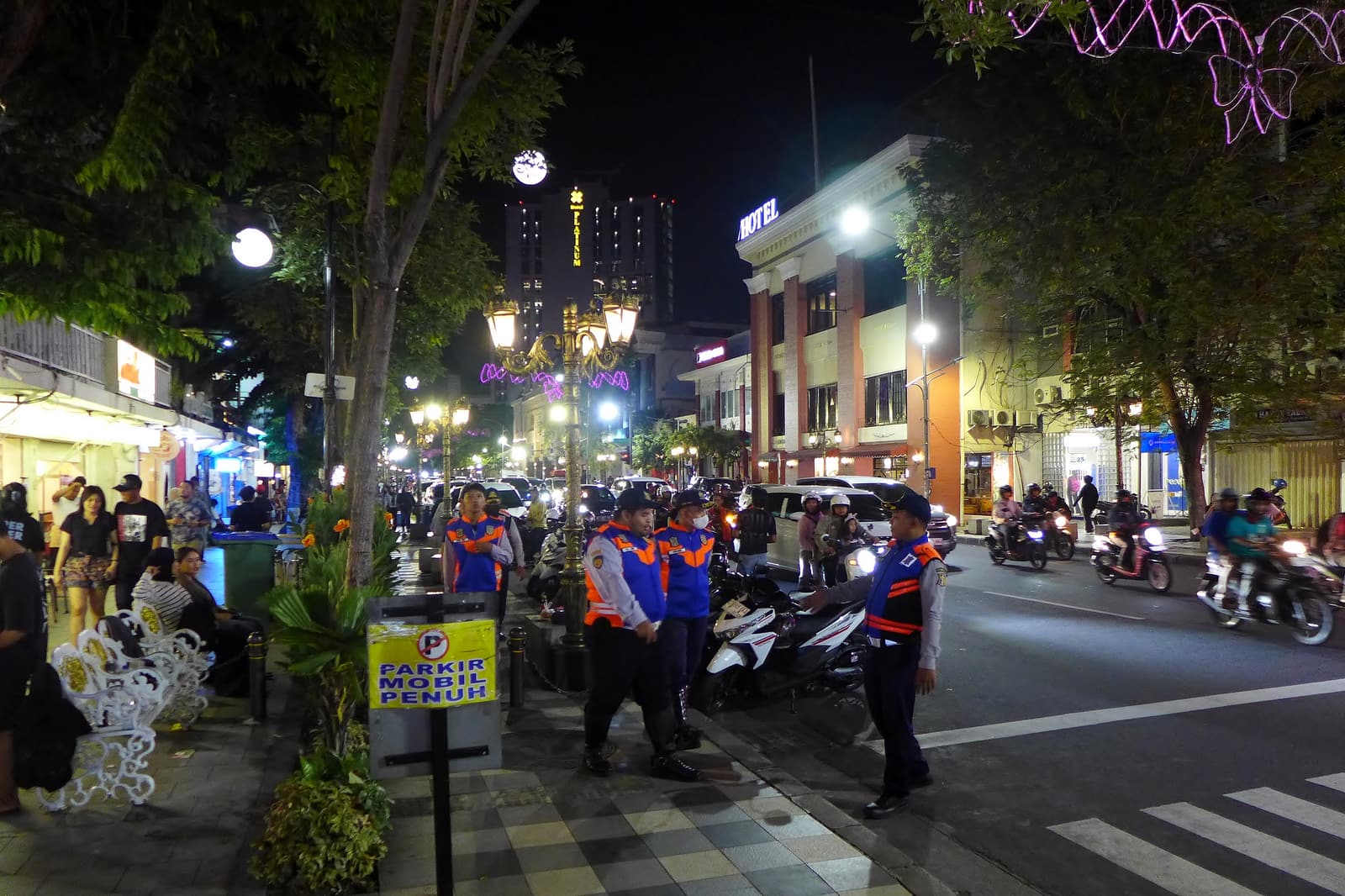 A February 2025 night on Jalan Tunjungan, with parking attendants and motorcycles crowding the pavement edge, cars in traffic, illuminated facades and a vertical PLATINUM sign in the distance.