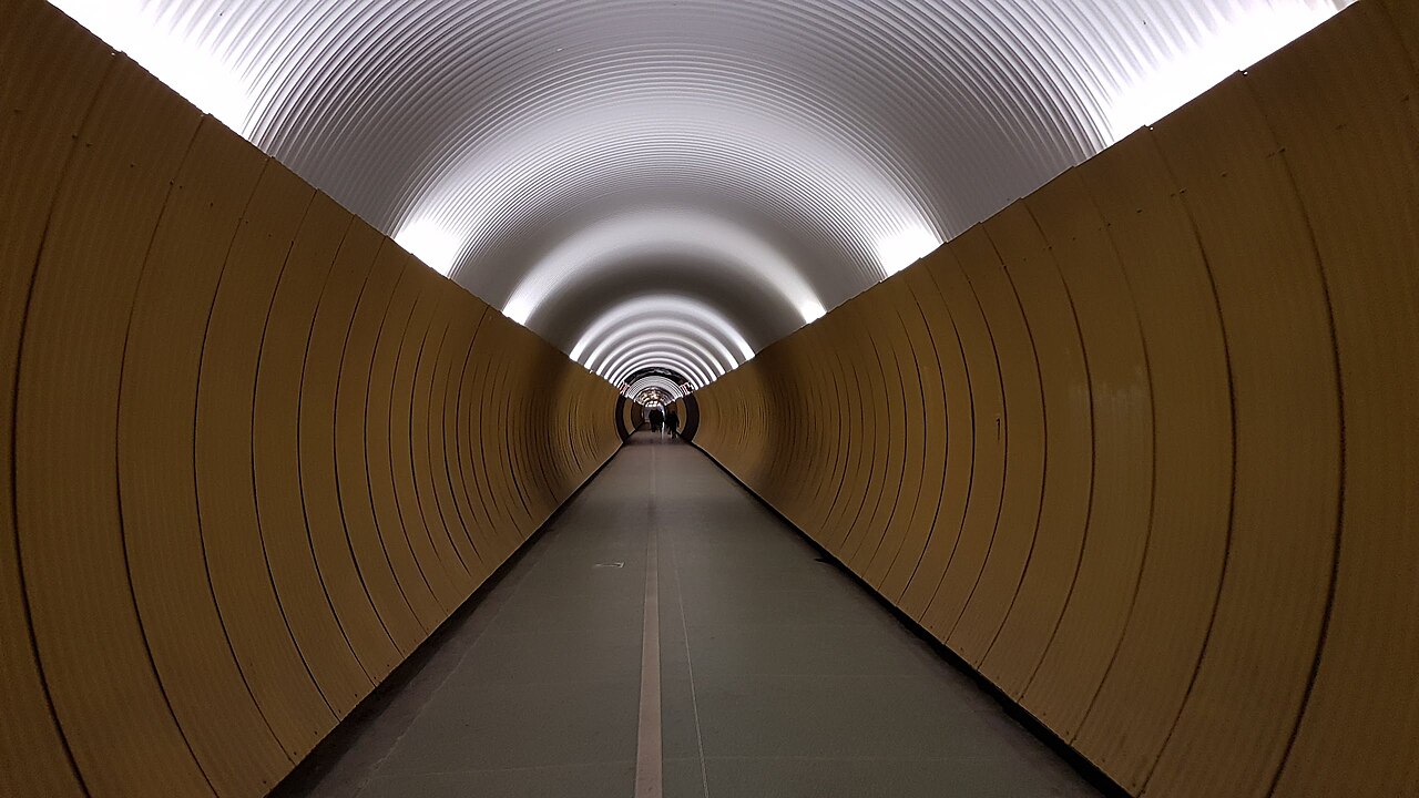 Yellow wall panels and overhead lights recede through Stockholm’s Brunkeberg Tunnel, with separate pedestrian and cycle markings on the floor.