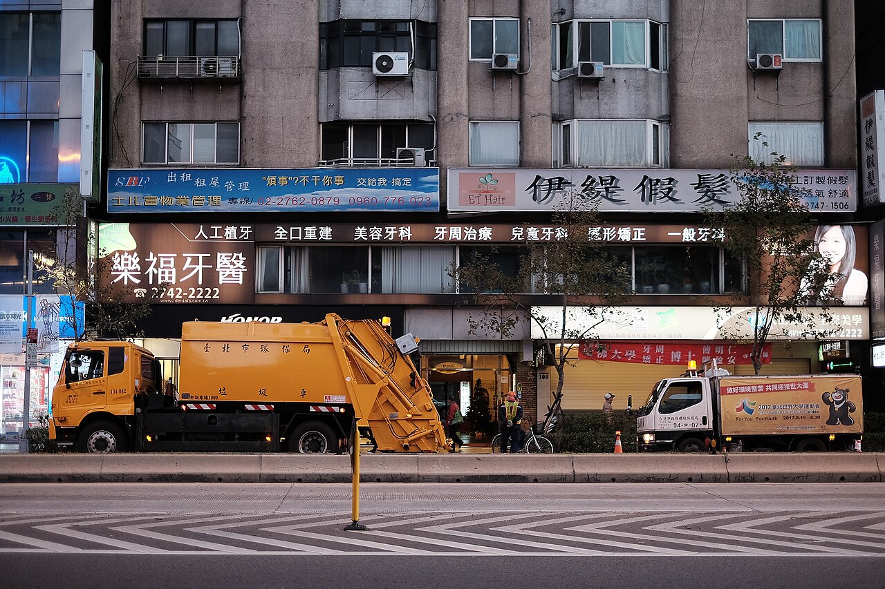 黄昏时分，台北松山区一辆黄色垃圾车与一辆白色资源回收车并排停在商铺和住宅楼外。