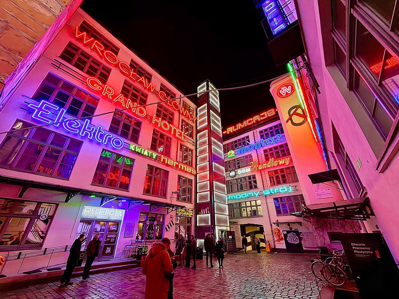 Historic neon signs in red, blue, green and white light cover the facades of Wrocław's Ruska 46 courtyard while visitors stand on the cobblestones below.