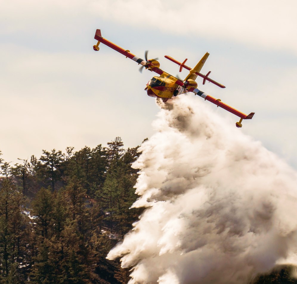 一架黄红相间的 Bridger Aerospace Super Scooper 汲水式灭火机在林木覆盖的山坡上方洒水。