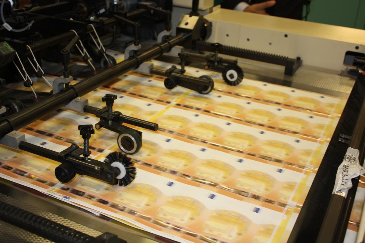 Sheets of euro banknotes passing beneath rollers on a production machine.