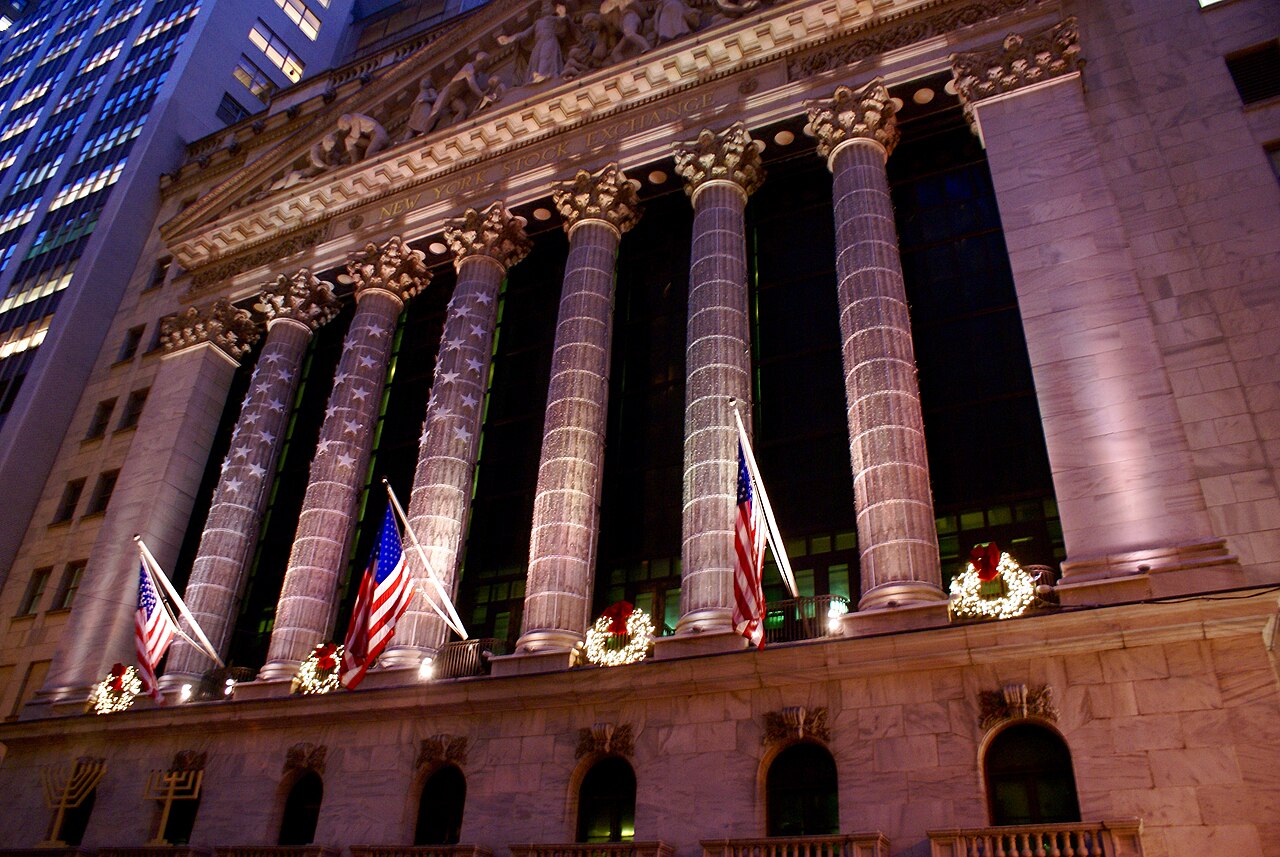 The columned New York Stock Exchange facade illuminated at night, with American flags above Broad Street.