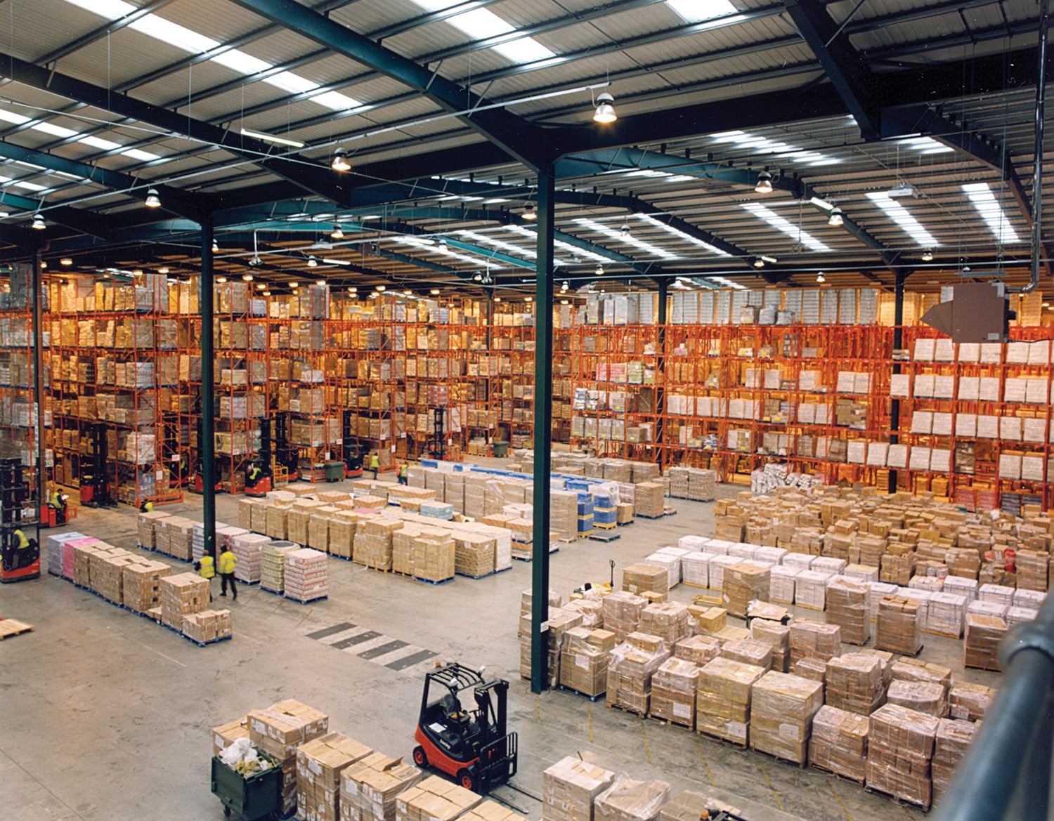 Workers and forklifts move among cartons and wrapped pallets beneath high warehouse racks.