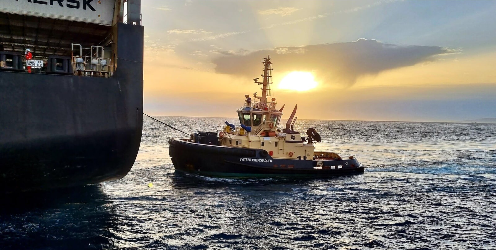夕阳低照，Svitzer Chefchaouen 号拖轮在一艘大型商船的船尾旁作业。