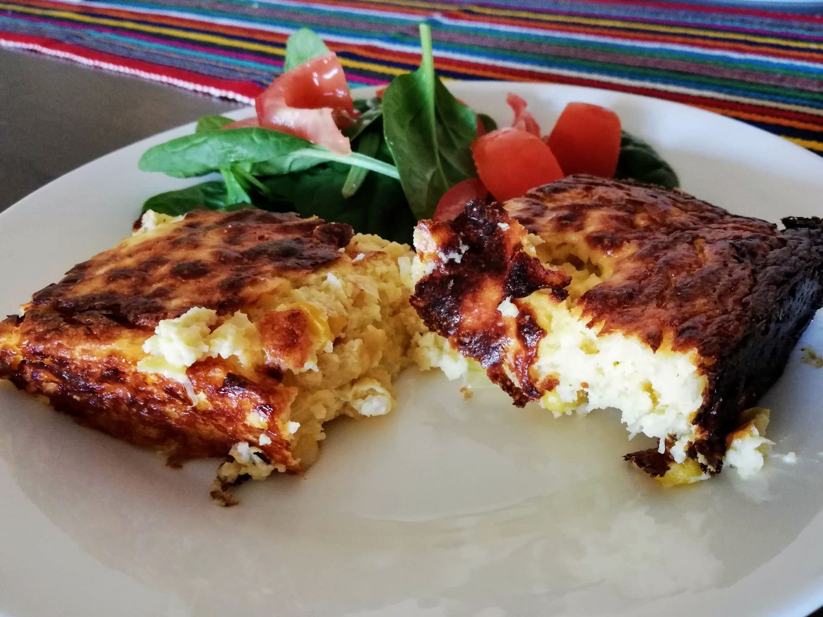Two browned portions of chipa guasu with exposed yellow corn and white cheese, served beside tomato and leafy salad on a white plate.