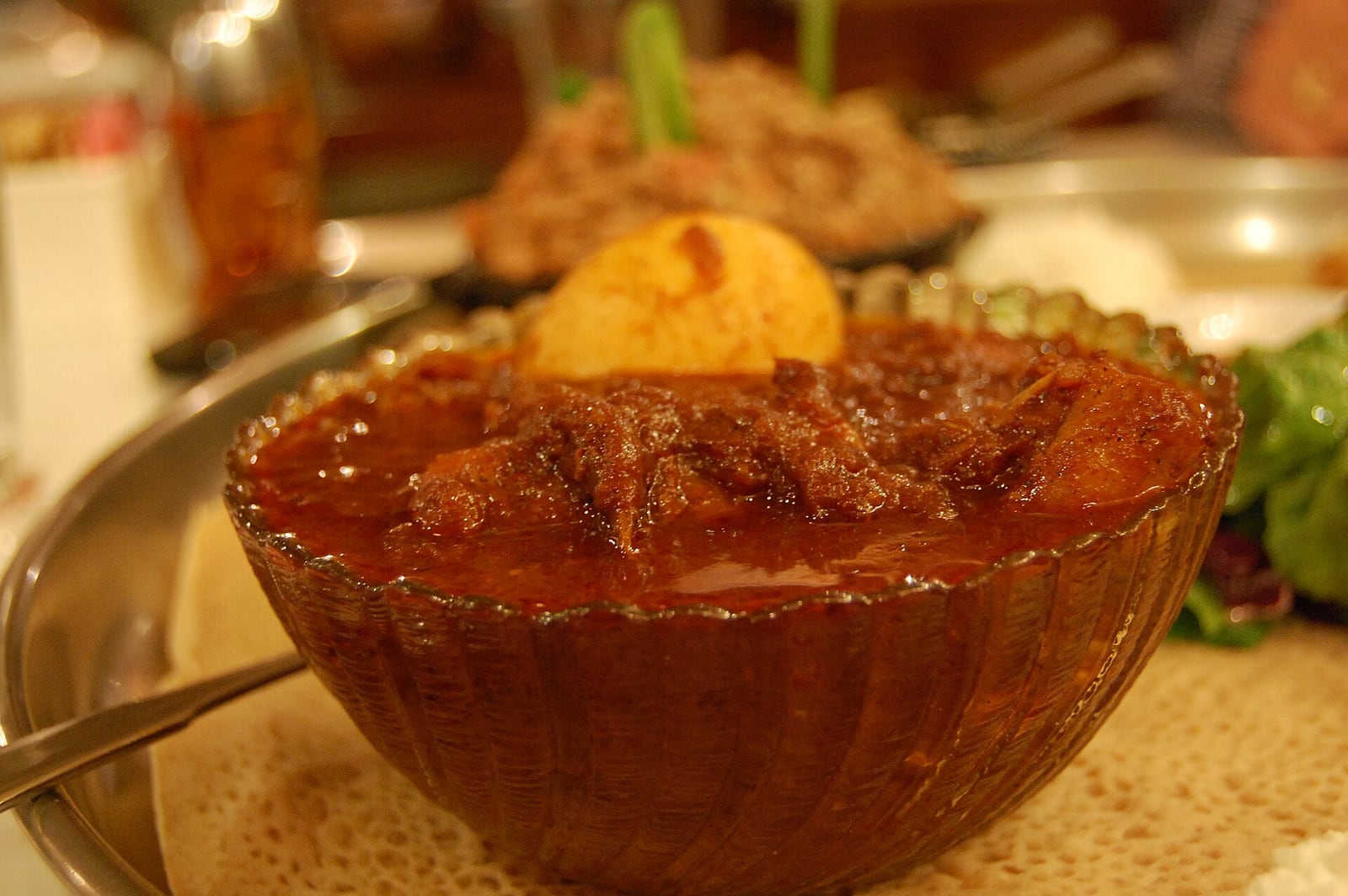 A glass bowl of brick-red doro wat with pieces of chicken and a boiled egg, set on pale, porous injera in a warmly lit restaurant.