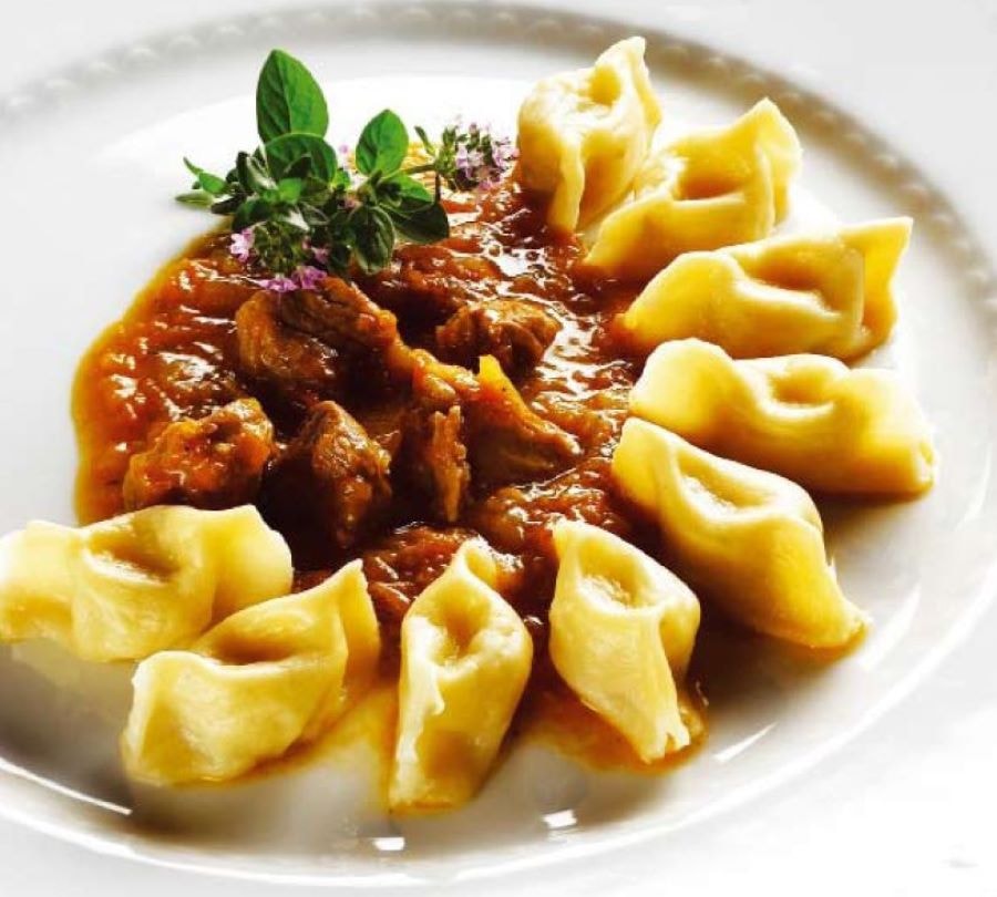 Golden, hat-shaped Idrija žlikrofi arranged around a chunky meat sauce, with green herbs and purple flowers on a white plate.
