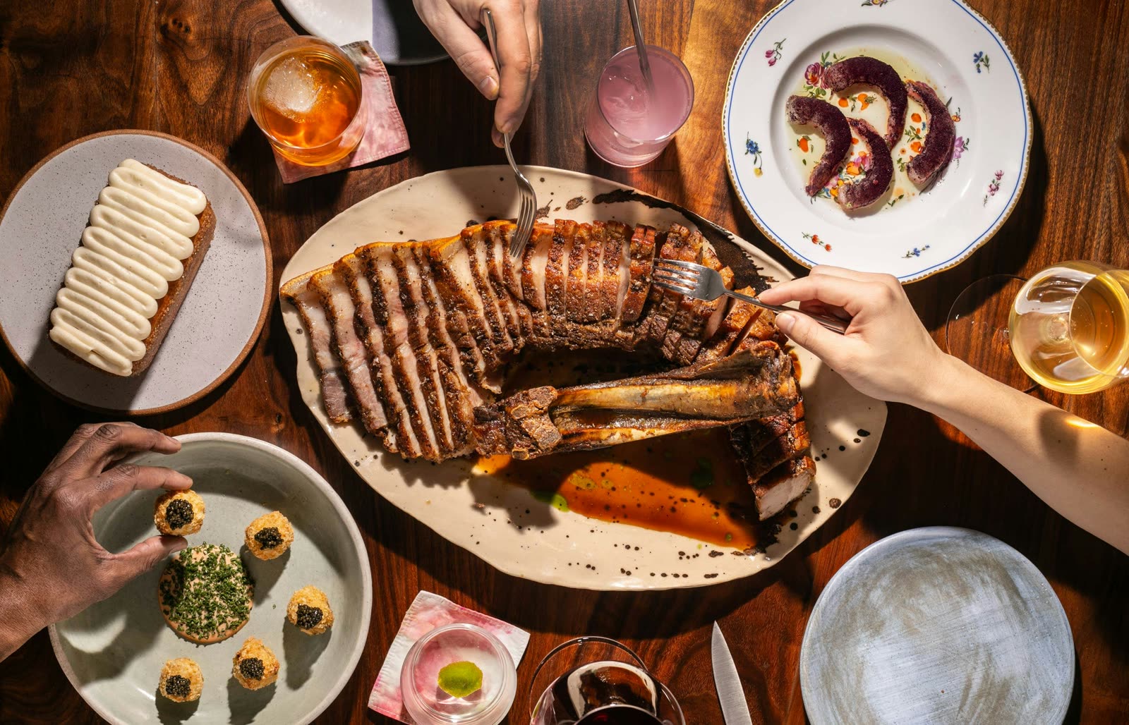 几位食客伸手越过一张木桌，桌上摆着 Kabawa 切片的 chuletas can can、小盘菜、葡萄酒与鸡尾酒。