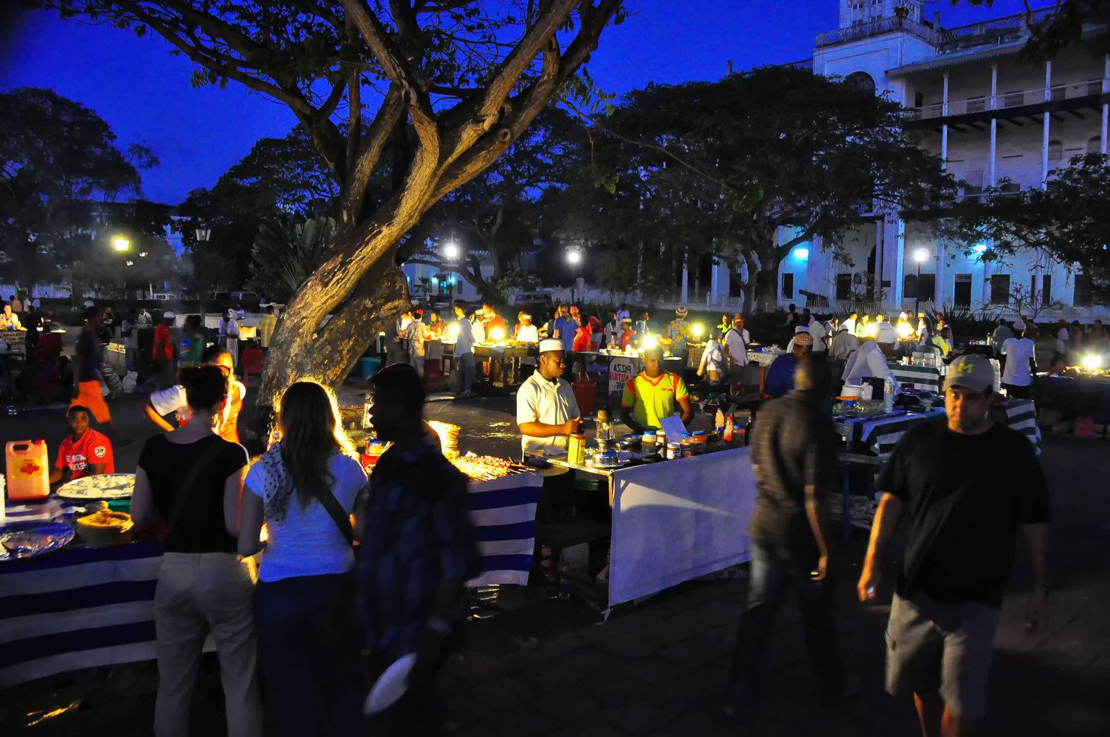 Food vendors and diners gather around illuminated market counters in Stone Town at blue hour.