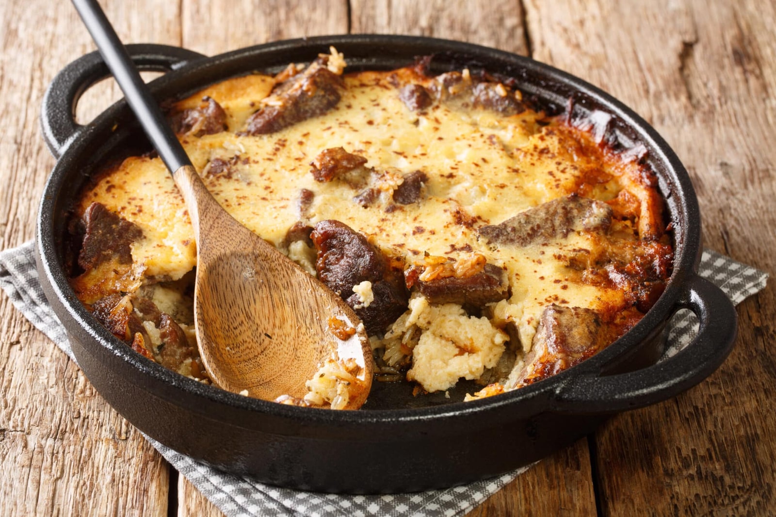 Lamb and rice beneath browned yogurt custard in a black casserole, with a wooden serving spoon.