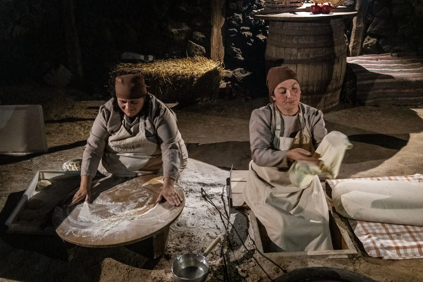 Two women make lavash by hand in Tsaghkunk's dim stone glkhatun, one rolling dough while the other stretches a thin sheet beside the tonir.
