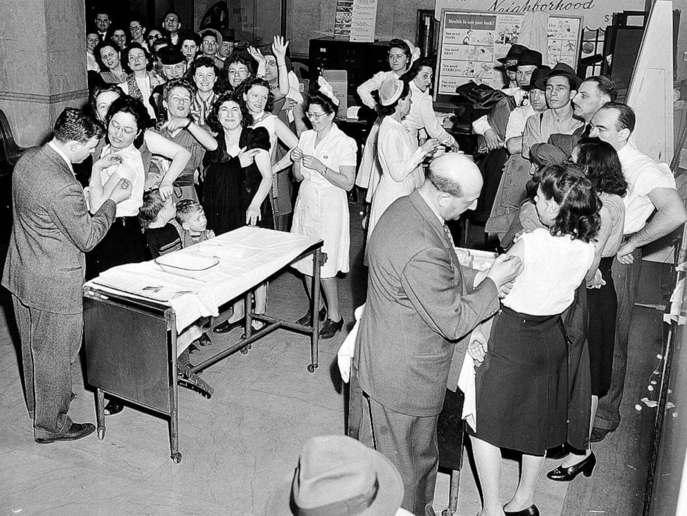 Doctors vaccinate two women while nurses and a crowd of adults and children fill a New York City Health Department room in 1947.
