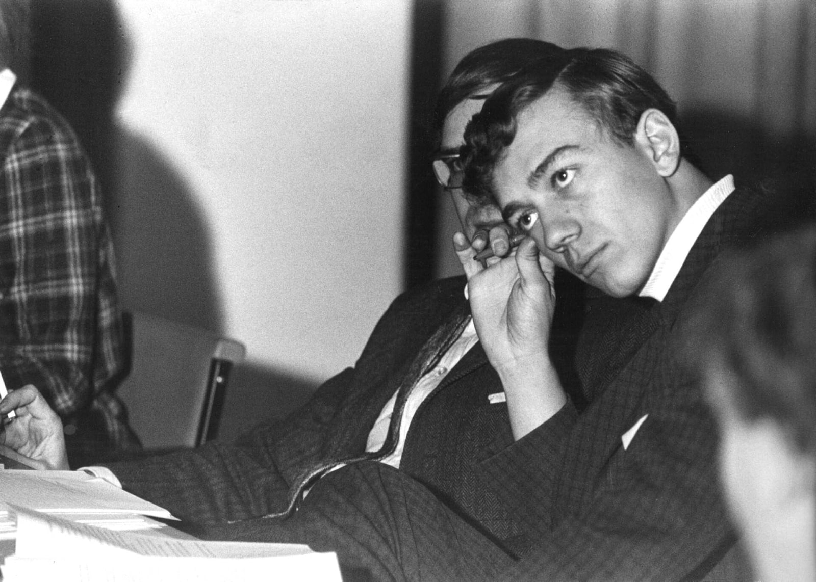 A young Pekka Puska leans his head on one hand while seated at a crowded meeting table in 1972.