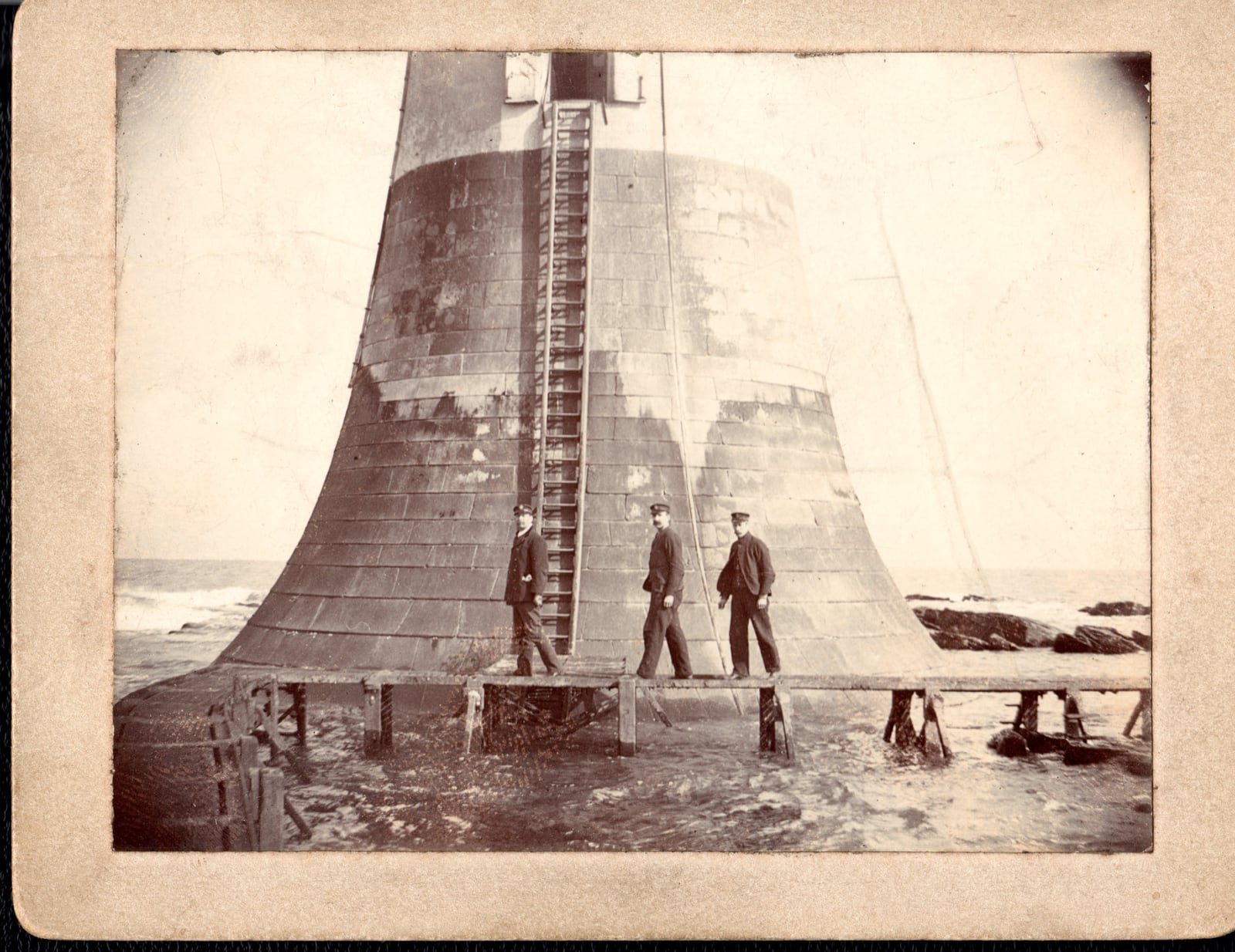 Three lighthouse keepers standing on a wooden landing platform beside the stone base of Bell Rock Lighthouse.