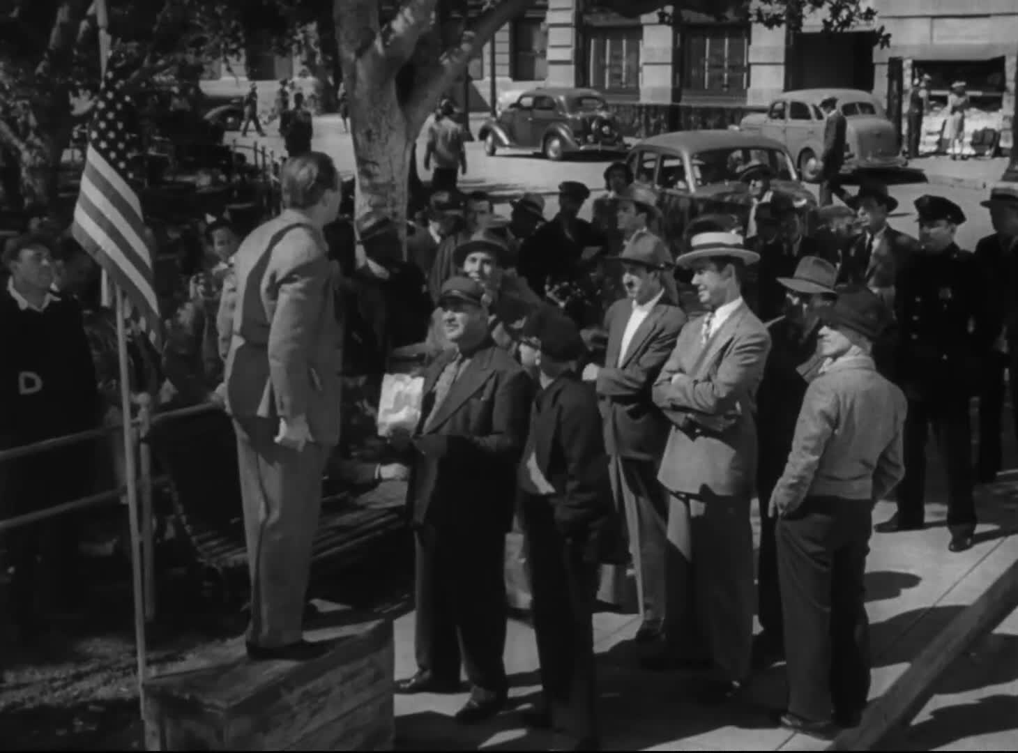 1945 年影片《别上当》中的黑白画面：一名街头演说者正在美国国旗旁向围拢的人群讲话。