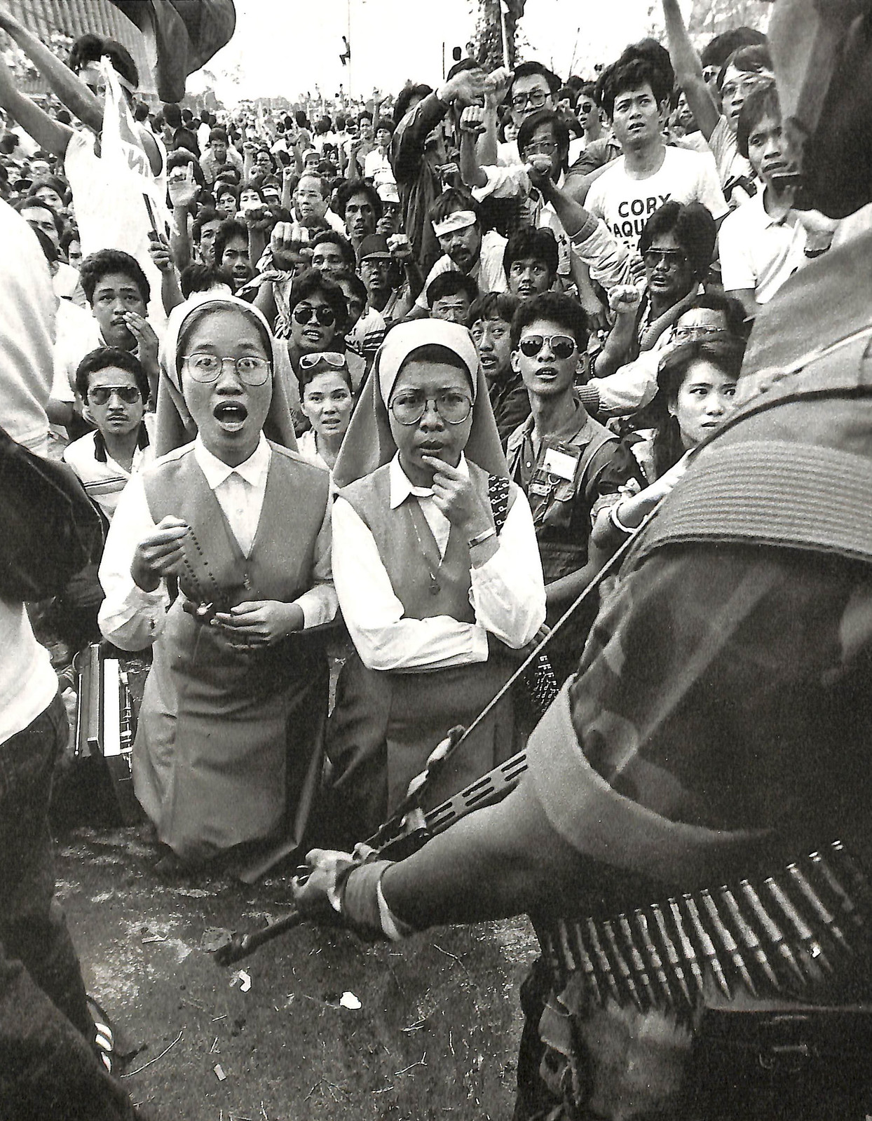 1986 年 EDSA 人民力量革命期间，两名天主教修女跪在一名持枪士兵面前，身后挤满围观人群。