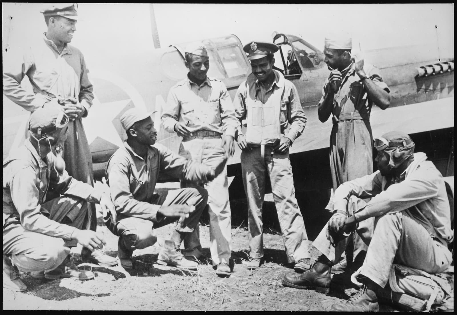 Seven Black U.S. Army Air Forces pilots gather beside a P-40 fighter at a Mediterranean air base in February 1944; several smile while one adjusts his flight gear.